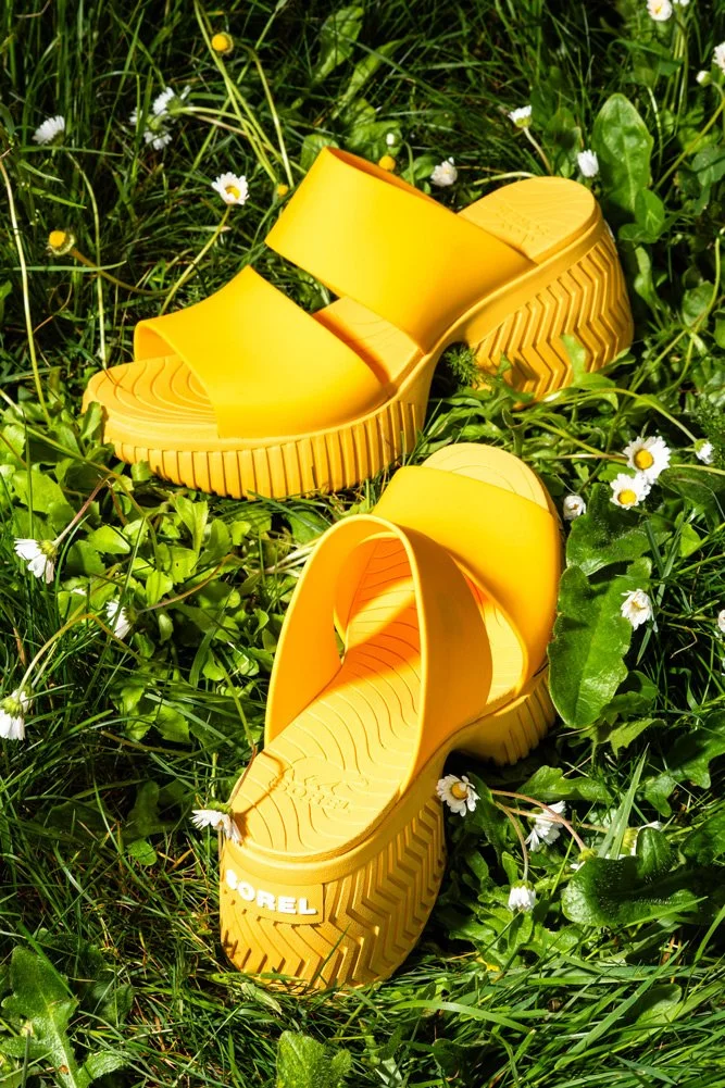A pair of bright yellow platform sandals lying on green grass surrounded by small white flowers.