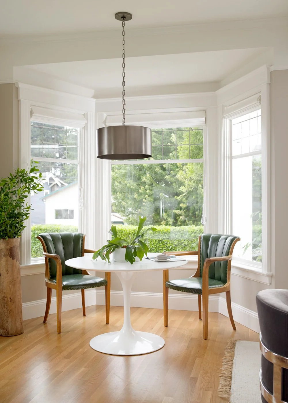A bright dining nook with a white round table, two green upholstered chairs, a large potted plant in the center, and large windows with greenery outside. A chandelier hangs above the table.