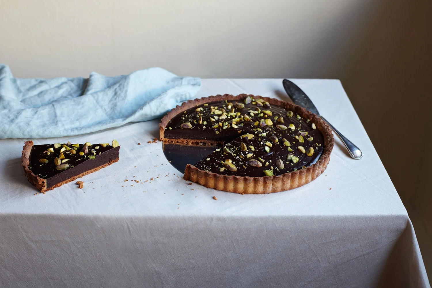 Chocolate tart with chopped pistachios on top, a slice removed, on a white tablecloth with a knife nearby, and a light blue cloth in the background.