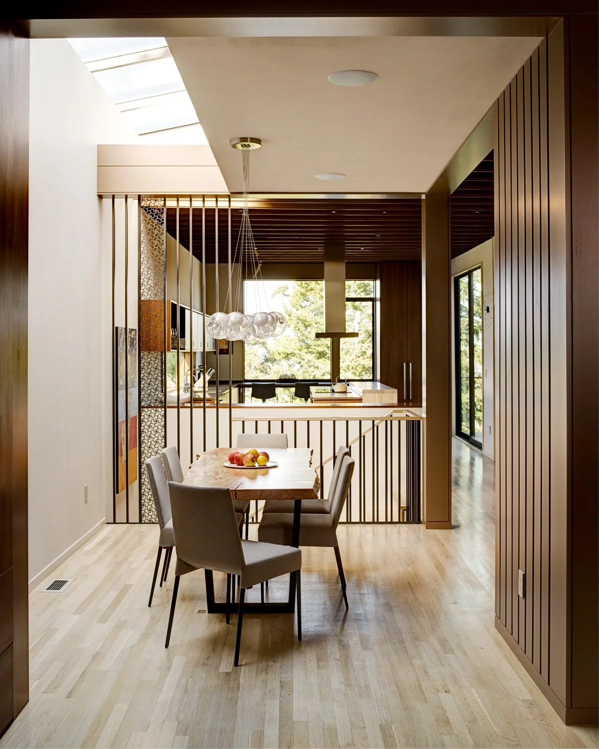 Modern dining area with wooden floors, a wooden table with six gray chairs, a fruit bowl on the table, and a chandelier with globe lights hanging above. Open space with large windows and natural light.