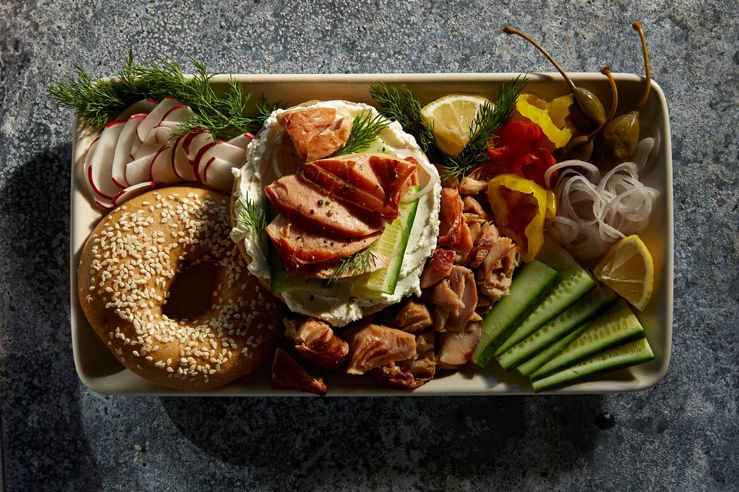 Platter of assorted appetizers including sliced radishes, sesame bagel, smoked salmon on cream cheese with herbs, cucumber slices, chopped chicken, and lemon wedges, garnished with fresh herbs.
