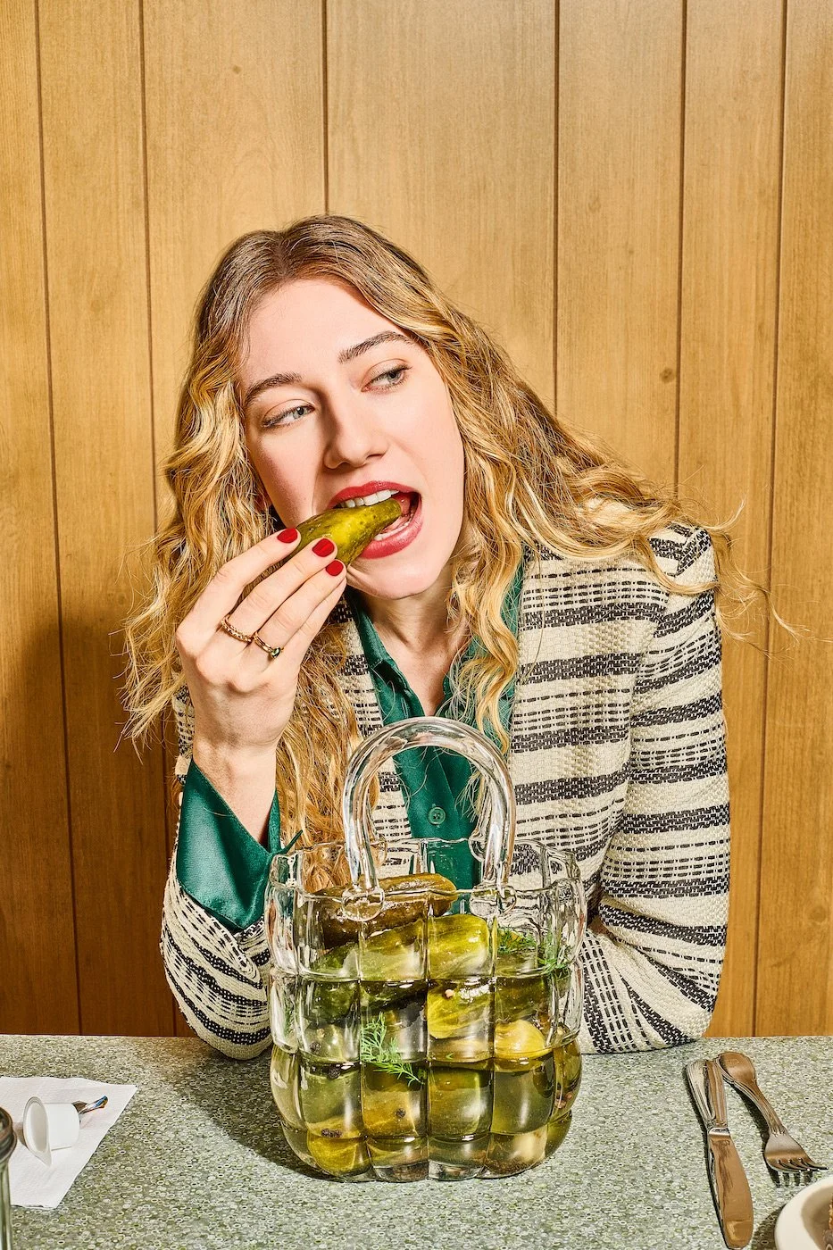Woman with curly blonde hair in a patterned blazer and green shirt eating a pickle from a glass container of pickles on a table.