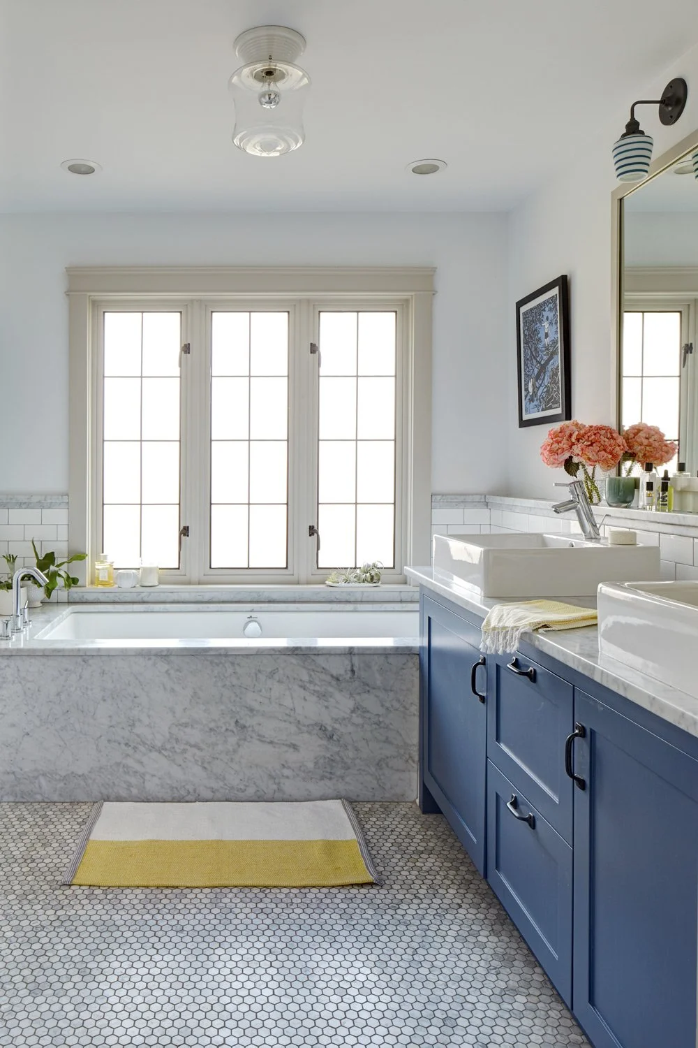 Bright bathroom with large window, marble bathtub, white and blue vanity, sink with pink flowers, framed artwork, and decorative toothbrush holder on the mirror