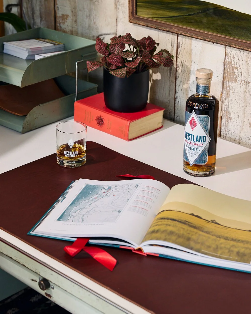 A desk with an open book showing a map and landscape photographs, a glass of whiskey, a bottle of whiskey, a closed red book, a black plant pot with a red-leafed plant, and a green metal tray with documents.