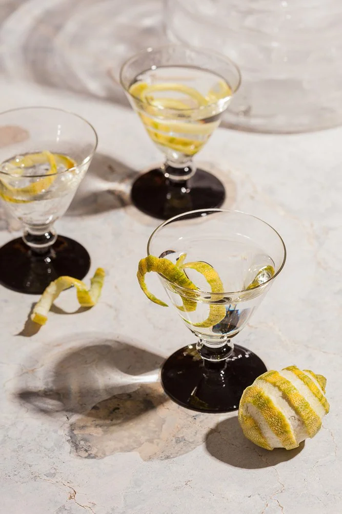 Three martini glasses with lemon twisted garnishes, filled with a clear drink, placed on a textured surface with shadows, and a decorative lemon-shaped object nearby.