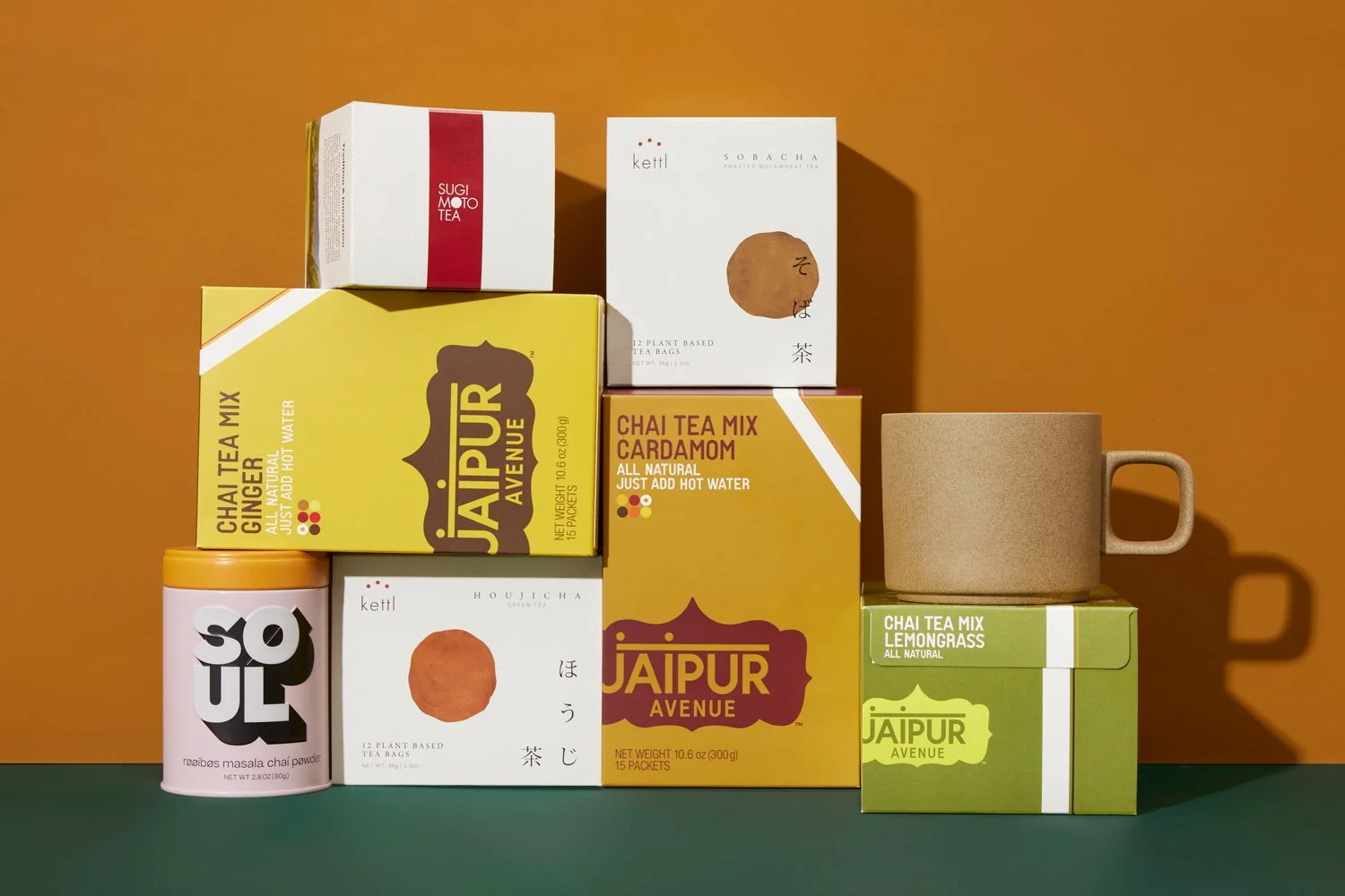 An assortment of tea boxes, a ceramic mug, and a container of masala chai powder, all stacked against a two-tone orange and green background.