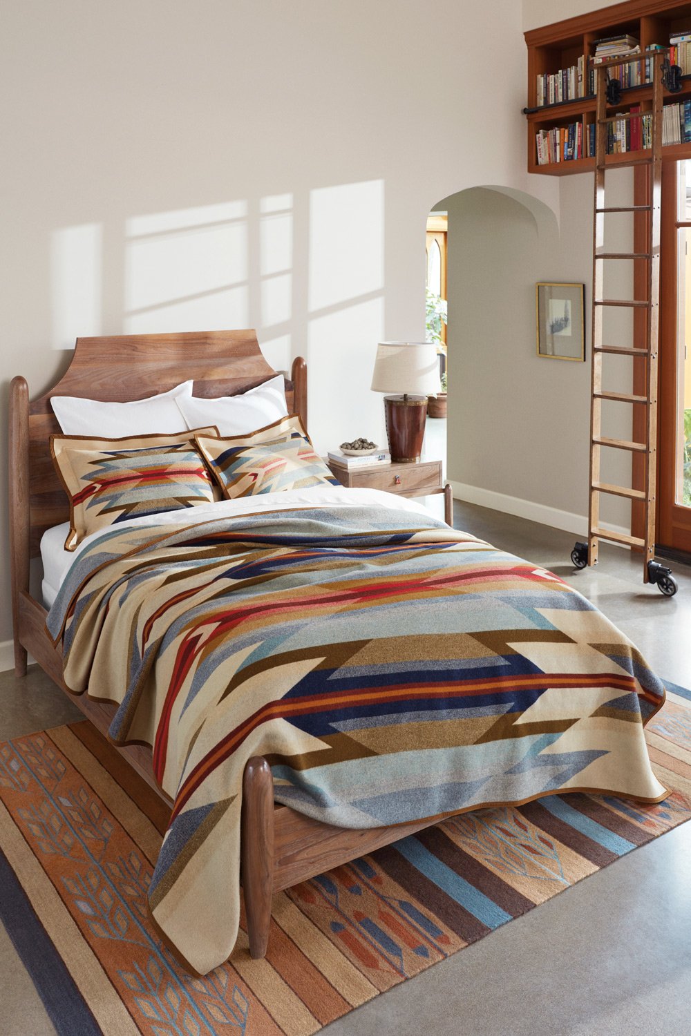 A bedroom with a wooden bed, a colorful southwestern pattern blanket and pillows, a wooden nightstand with a lamp, bookshelf on a rolling ladder, window with sunlight, and a framed picture on the wall.