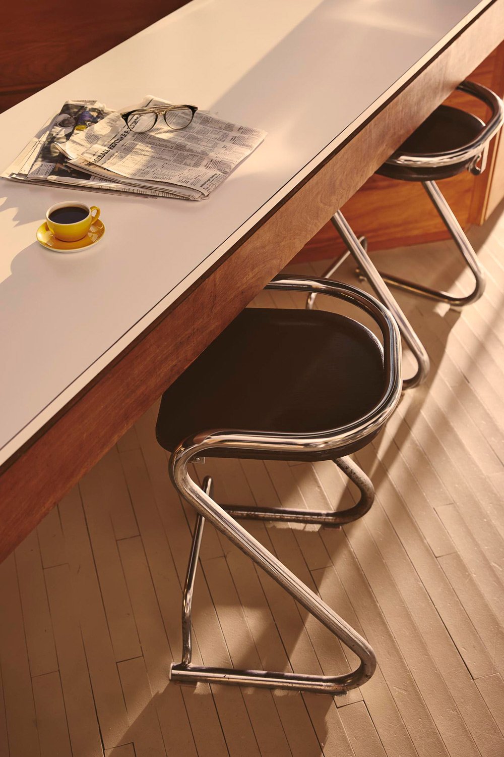 A breakfast bar with a yellow cup of coffee, a newspaper, and eyeglasses on top. Two black chairs with metal frames are positioned against the bar. The bar has a white top and wooden sides, and the floor is light wood.