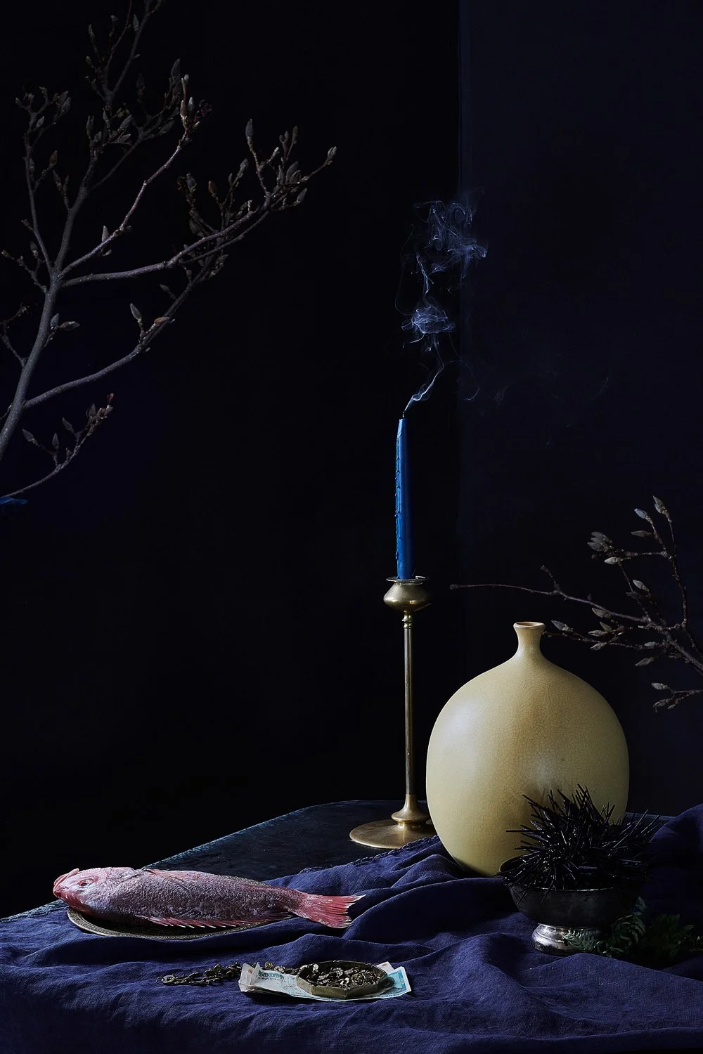 Still life with a fish, a blue candle in a candlestick with smoke, a beige vase, branches, jewelry, and a black decorative object, all arranged on a dark cloth, against a dark background.
