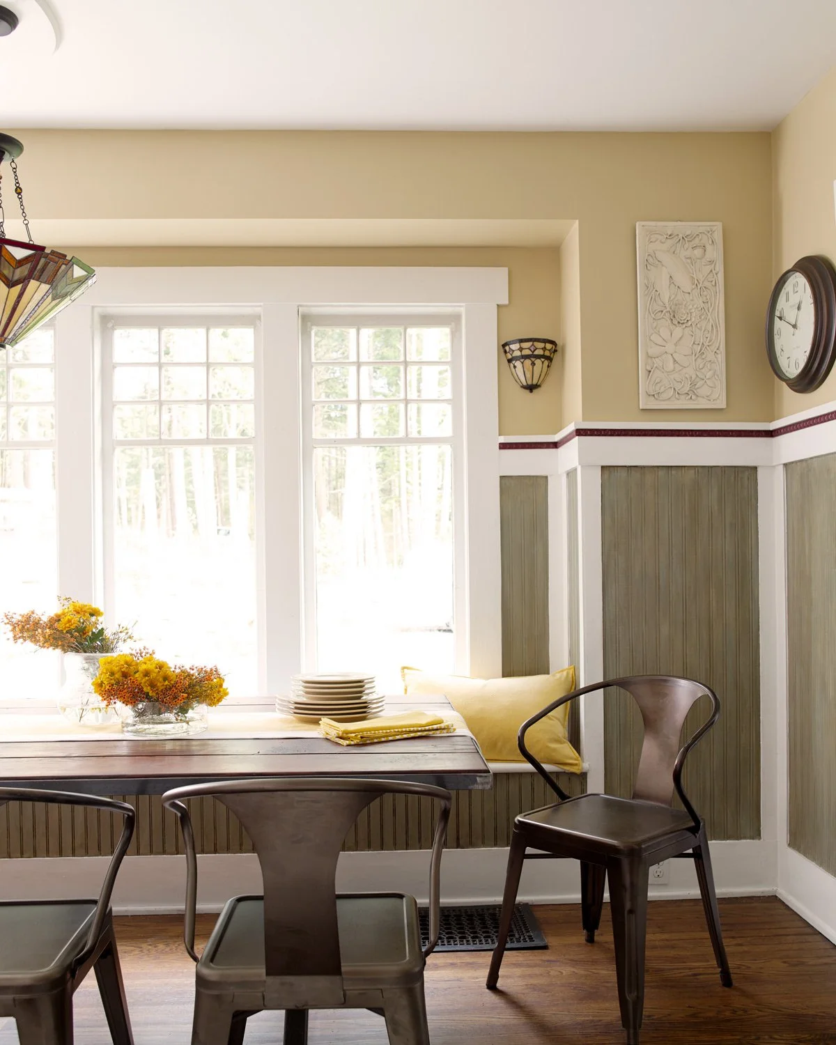 A dining nook with a table set next to large windows, yellow flowers in a glass bowl, and stacked plates. Wooden chairs surround the table, with a yellow pillow on a window seat. A wall clock and decorative wall art are visible on the beige and green