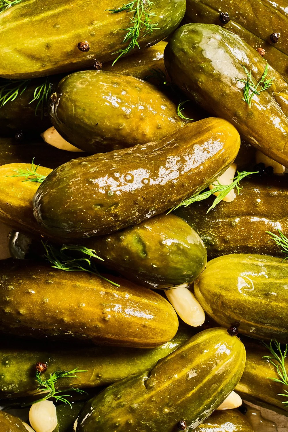 Close-up of green pickles with garlic cloves, black peppercorns, and sprigs of dill or herbs.