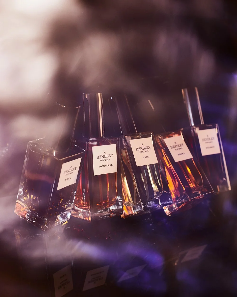 A collection of perfume bottles displayed against a dark background, with labels reading Hendley Perfumes and various scent names such as Rosenthal, Jade, Amour, and Moth. The bottles are angled and reflect light, creating a luxurious aesthetic.