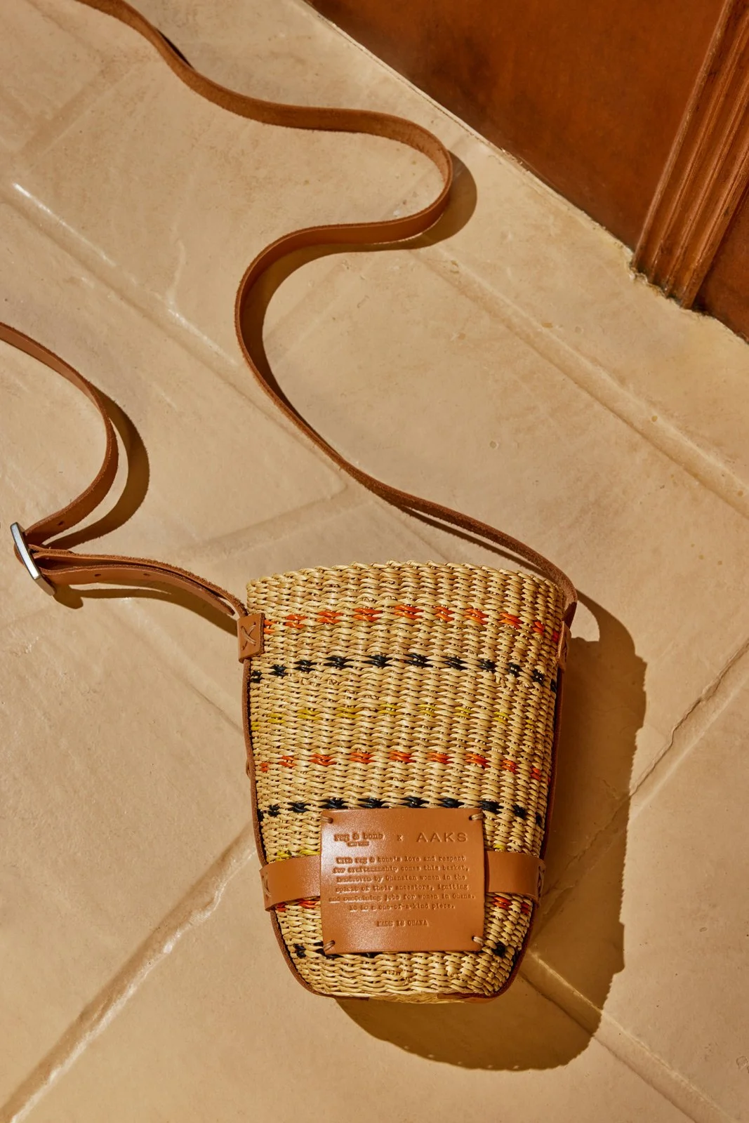 A woven straw crossbody bag with leather straps and details, placed on a tiled floor.