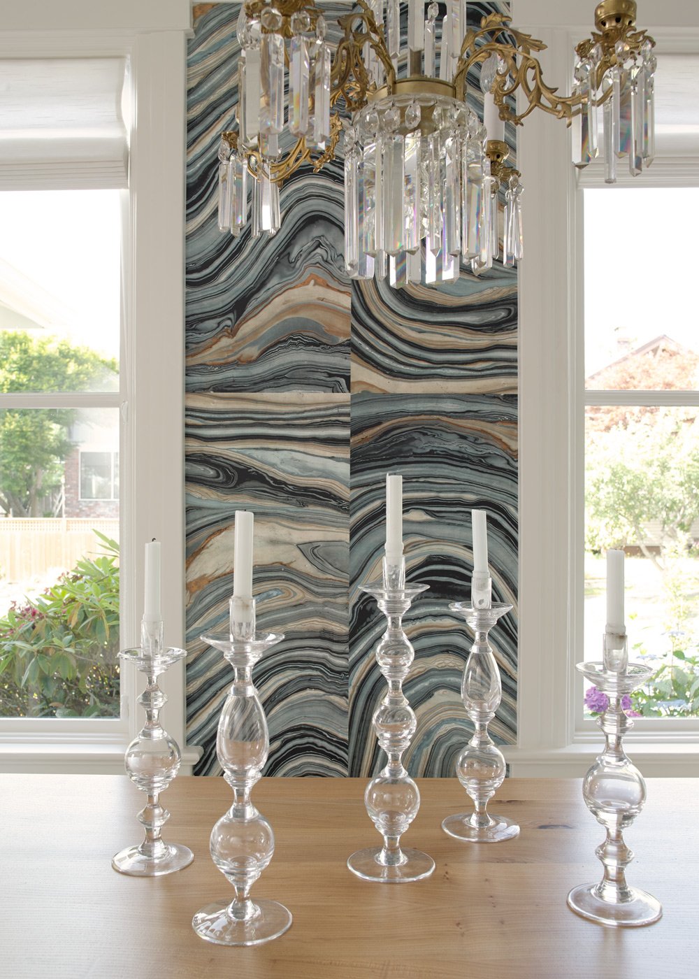 Chandelier with crystal prisms hanging above a table with five glass candle holders and white candles, in a room with large windows, a marble-patterned wall, and greenery outside.