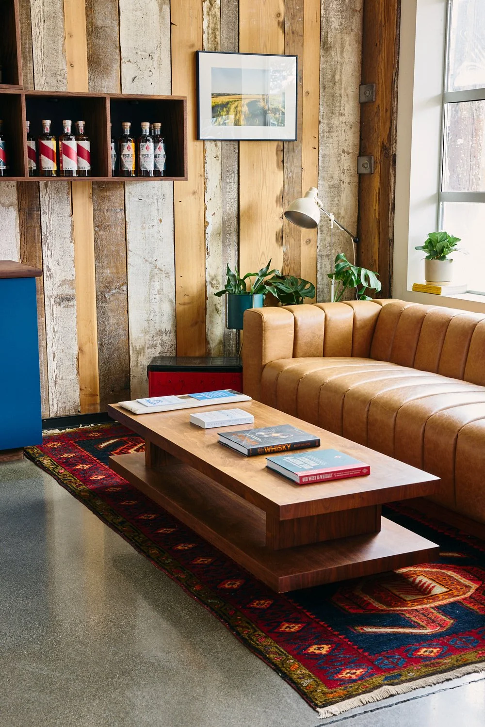Cozy lounge area with a brown leather sofa, wooden coffee table with magazines, a patterned rug, and potted plants near a window and wooden wall.