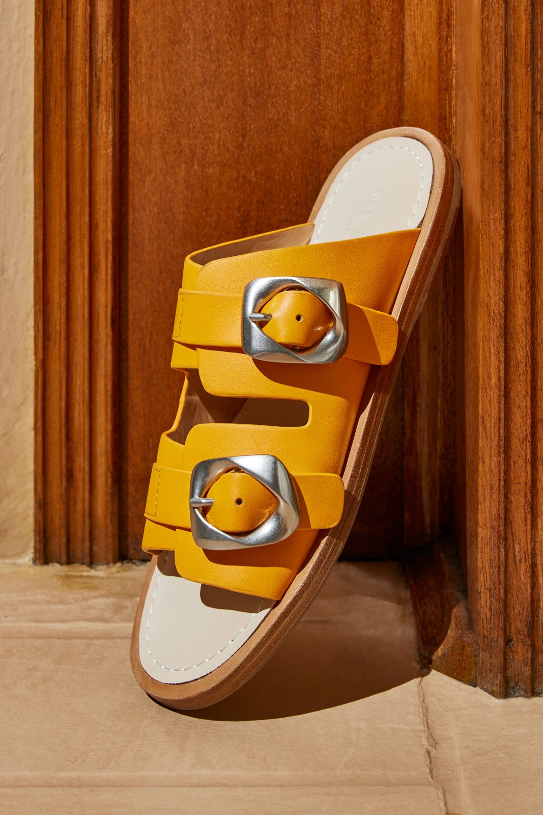 Yellow sandal with silver buckles, mounted on a wooden surface with a wooden background.