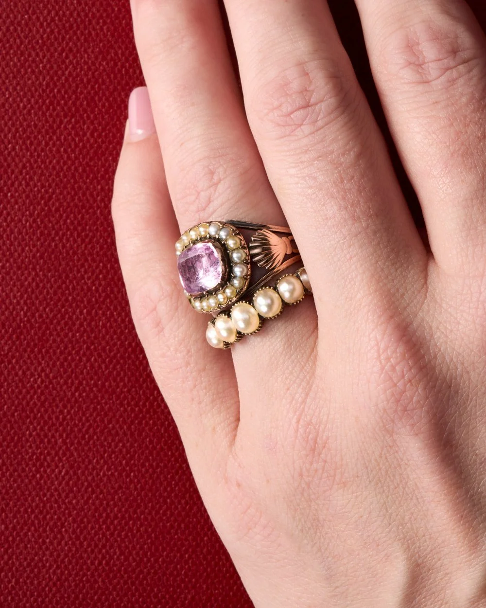 Close-up of a hand wearing a gold ring with a large pink gemstone surrounded by small pearls, and a band of pearls on the side, against a red textured background.