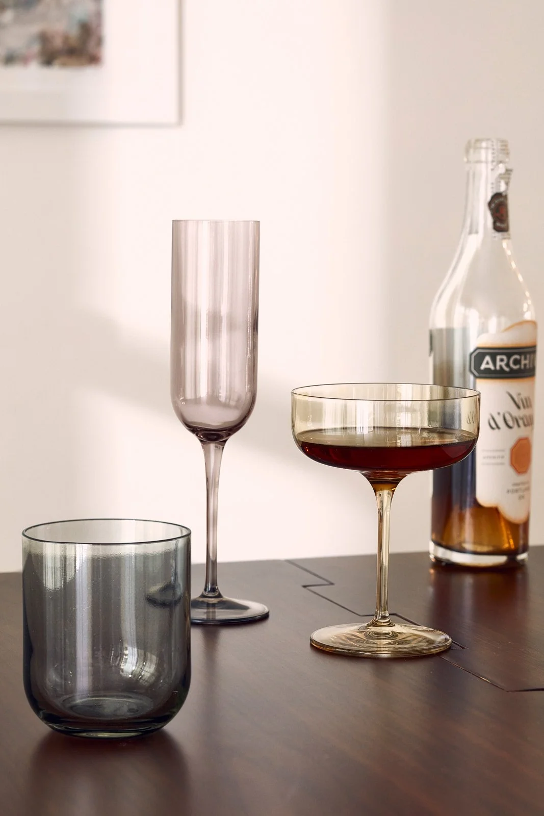 A clear glass tumbler, a purple-tinted champagne flute, a gold-rimmed wine glass filled with red wine, and a bottle of alcohol on a dark wooden table.