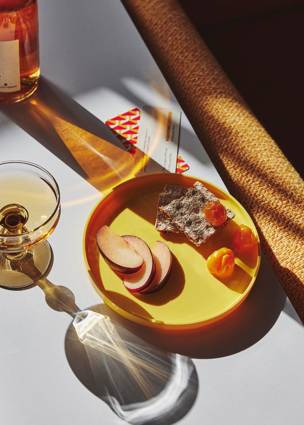 A yellow plate with sliced pears, a piece of cake dusted with powdered sugar and topped with orange jam, and two orange dollops of cream, positioned next to a glass of white wine on a white table.