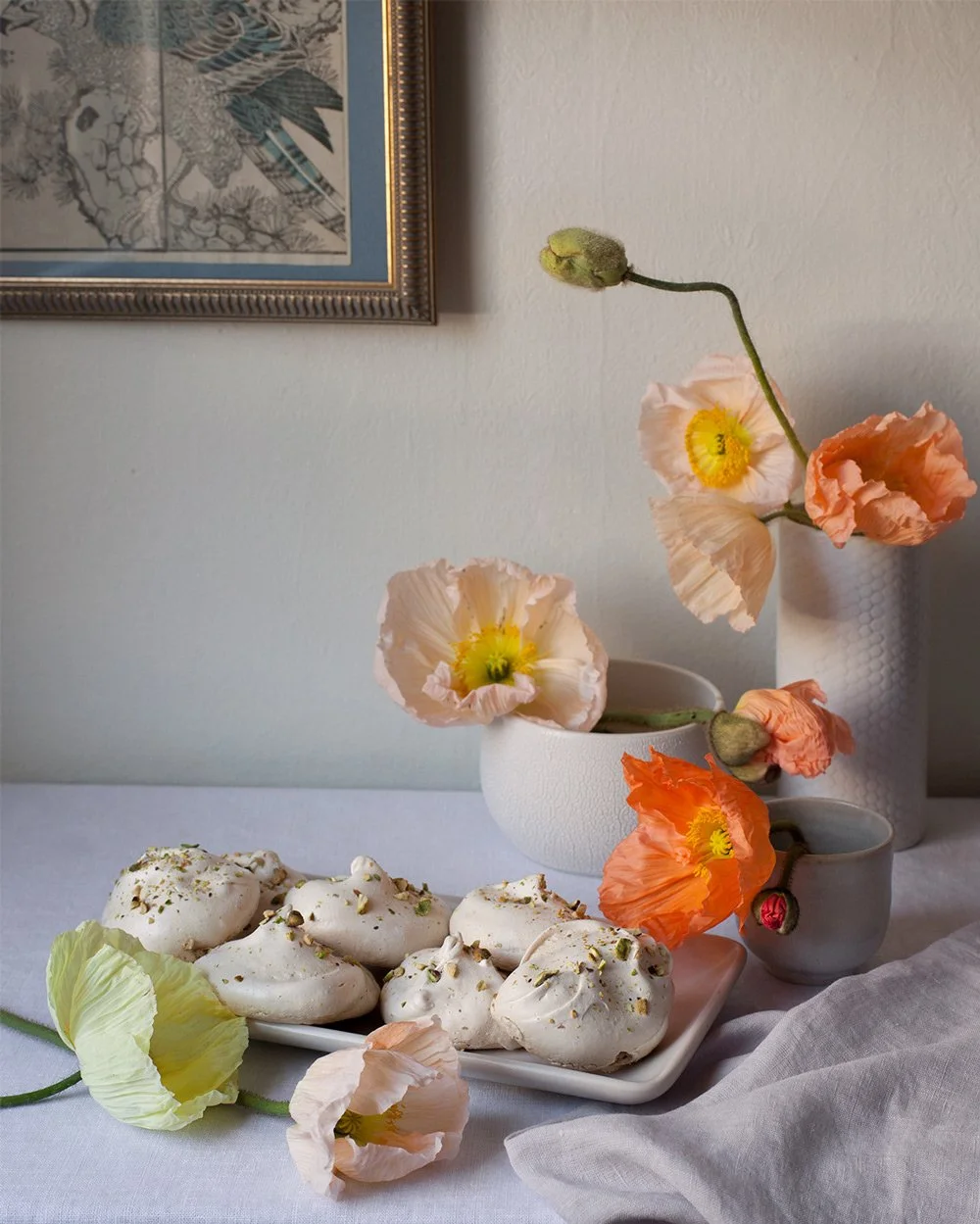 A table with a plate of white meringue cookies sprinkled with chopped pistachios, and a arrangement of peach, orange, and cream poppy flowers in white vases against a neutral wall and a framed artwork.