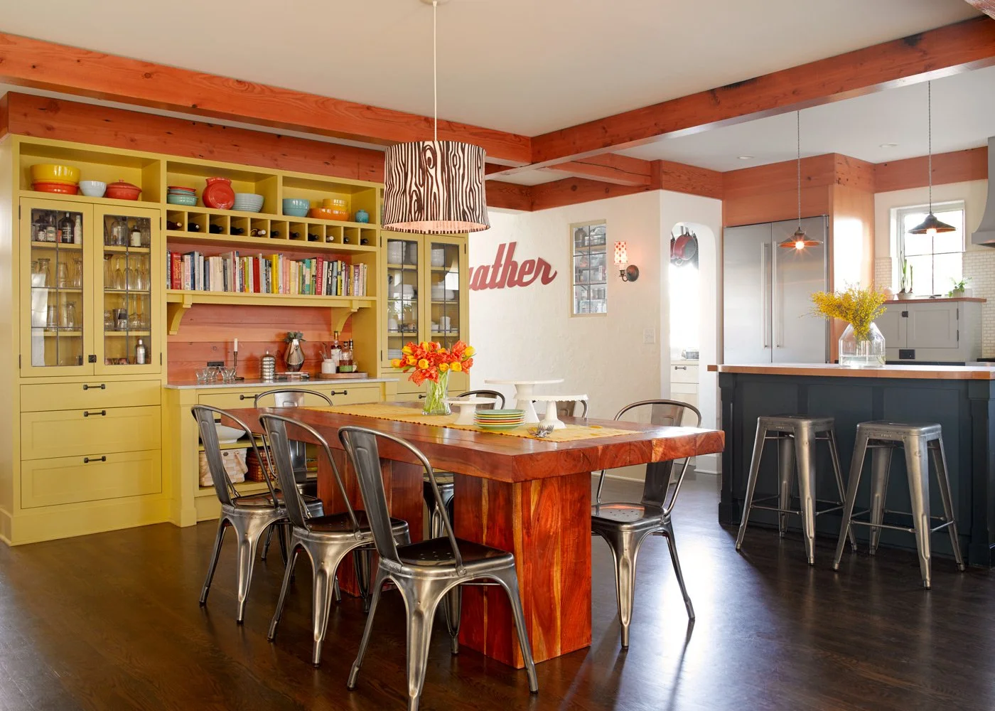 A bright, modern kitchen and dining area with yellow cabinets, a large wooden dining table with metal chairs, a kitchen island with barstools, and decorative elements like flowers and wall art.