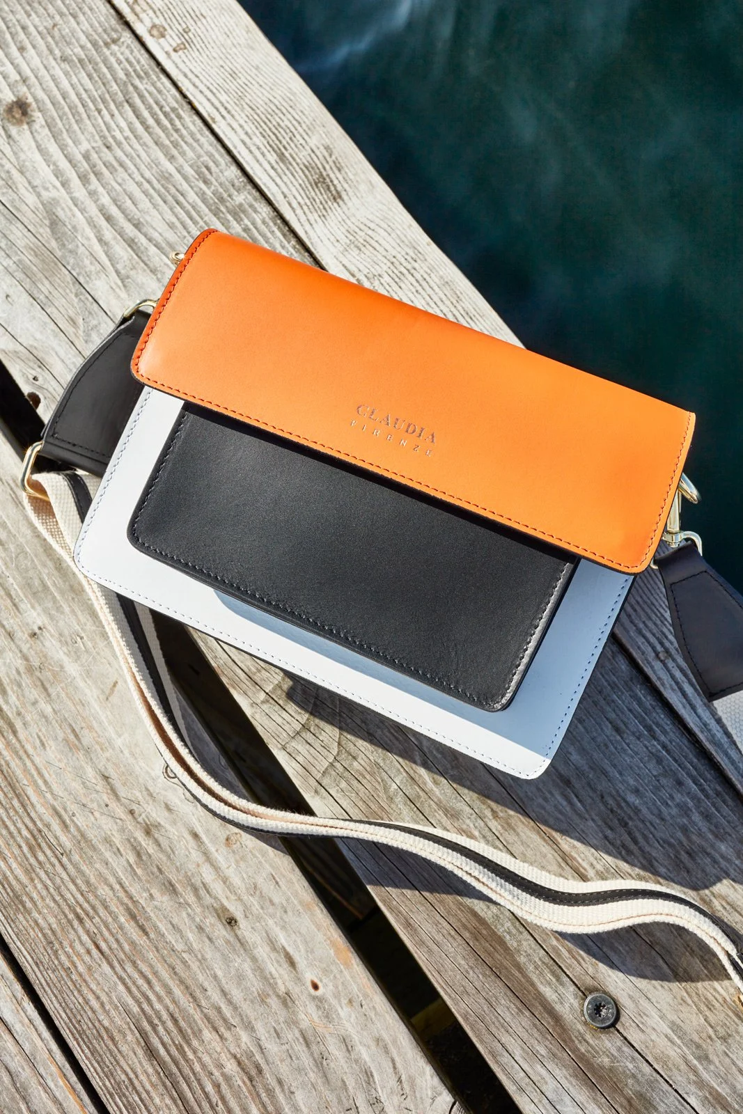 Colorful purses stacked on a wooden dock near the water, with a black, white, and orange handbag with a strap.