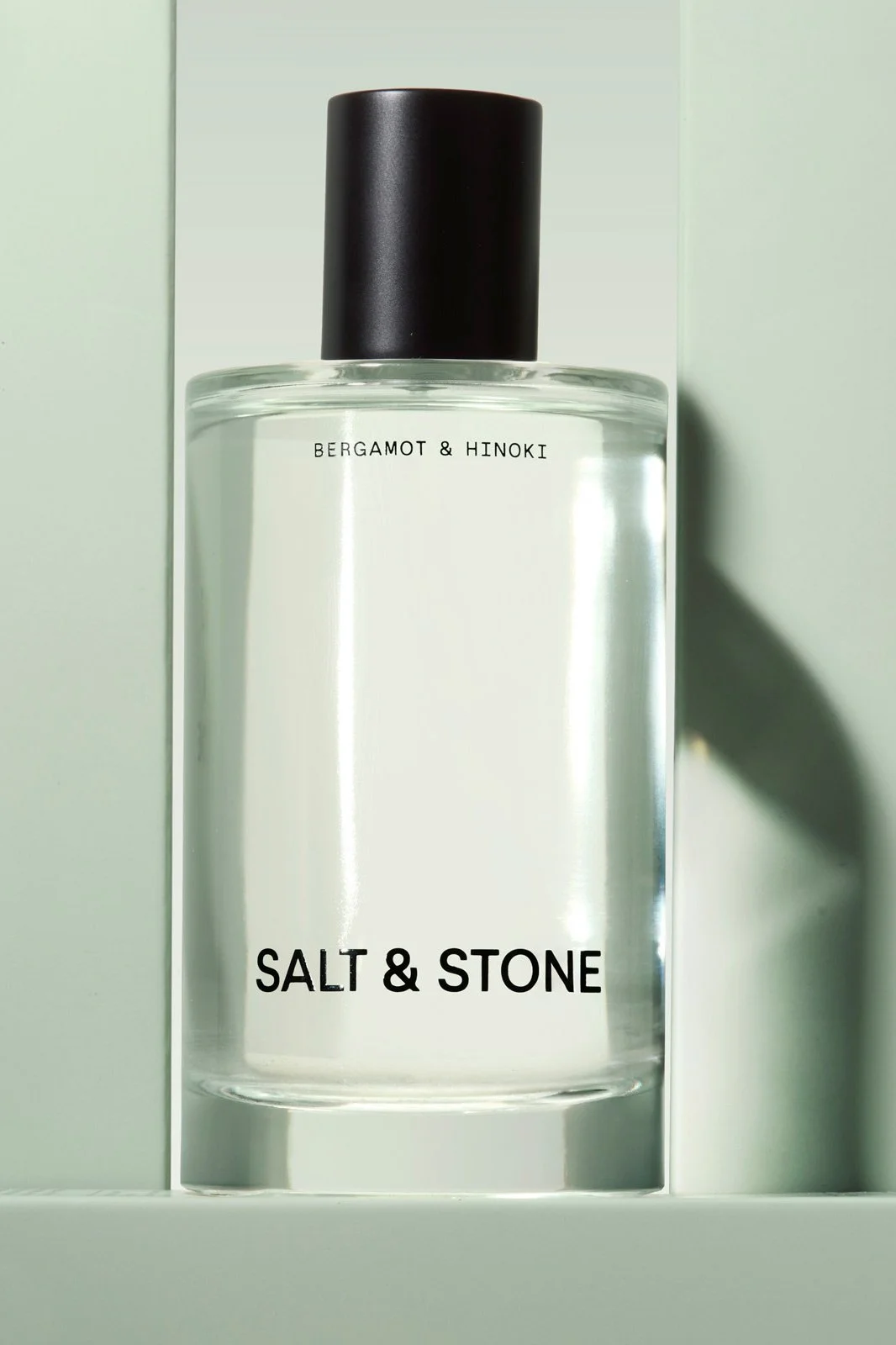 A clear glass bottle of Bergamot & Hinoki Salt & Stone fragrance with a black cap on a light background.