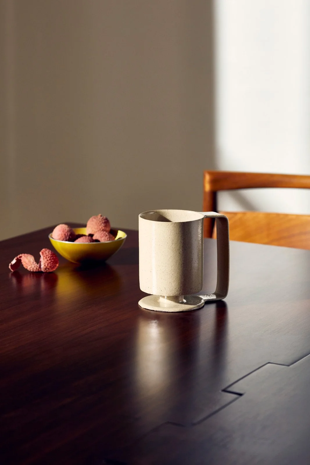 A ceramic mug and a bowl of lychee fruit on a wooden table, with a wooden chair in the background and soft natural lighting.