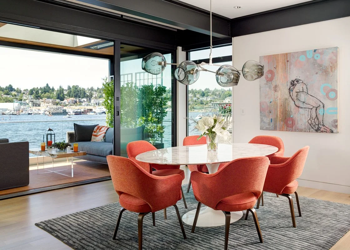 Modern dining area with a round marble table and six coral-colored chairs, a glass pendant light fixture, and a large window overlooking a water scene with boats and greenery.