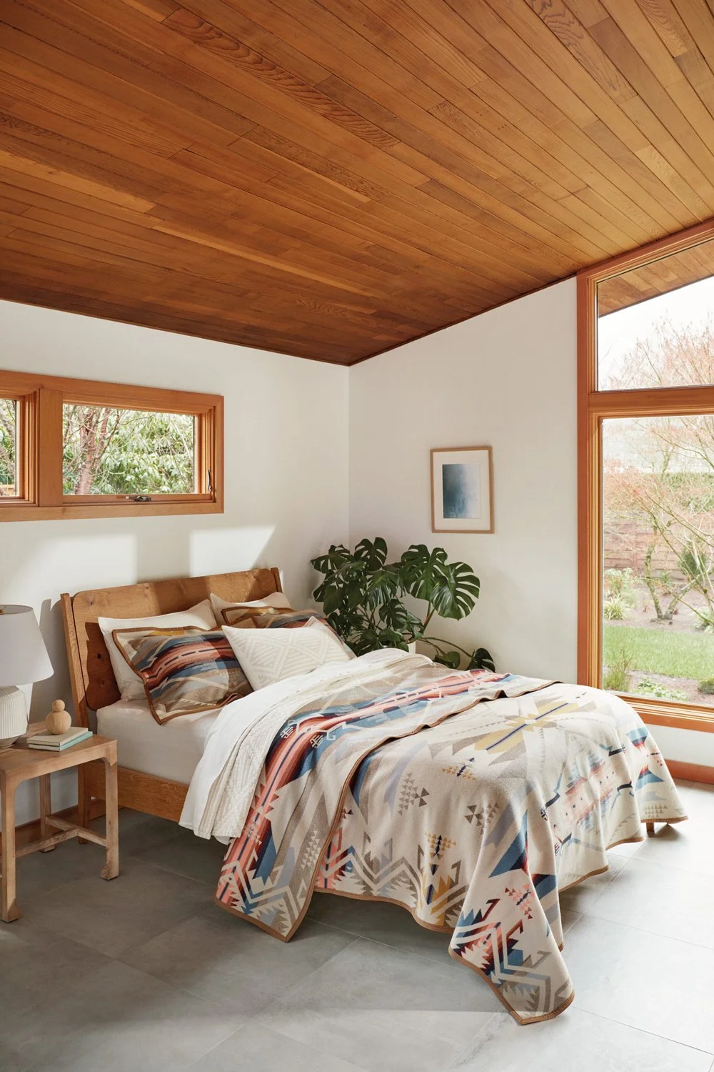 A bedroom with a wooden bed frame, patterned bedding, a small wooden nightstand with a lamp and books, and large windows showing an outdoor garden with trees and grass.