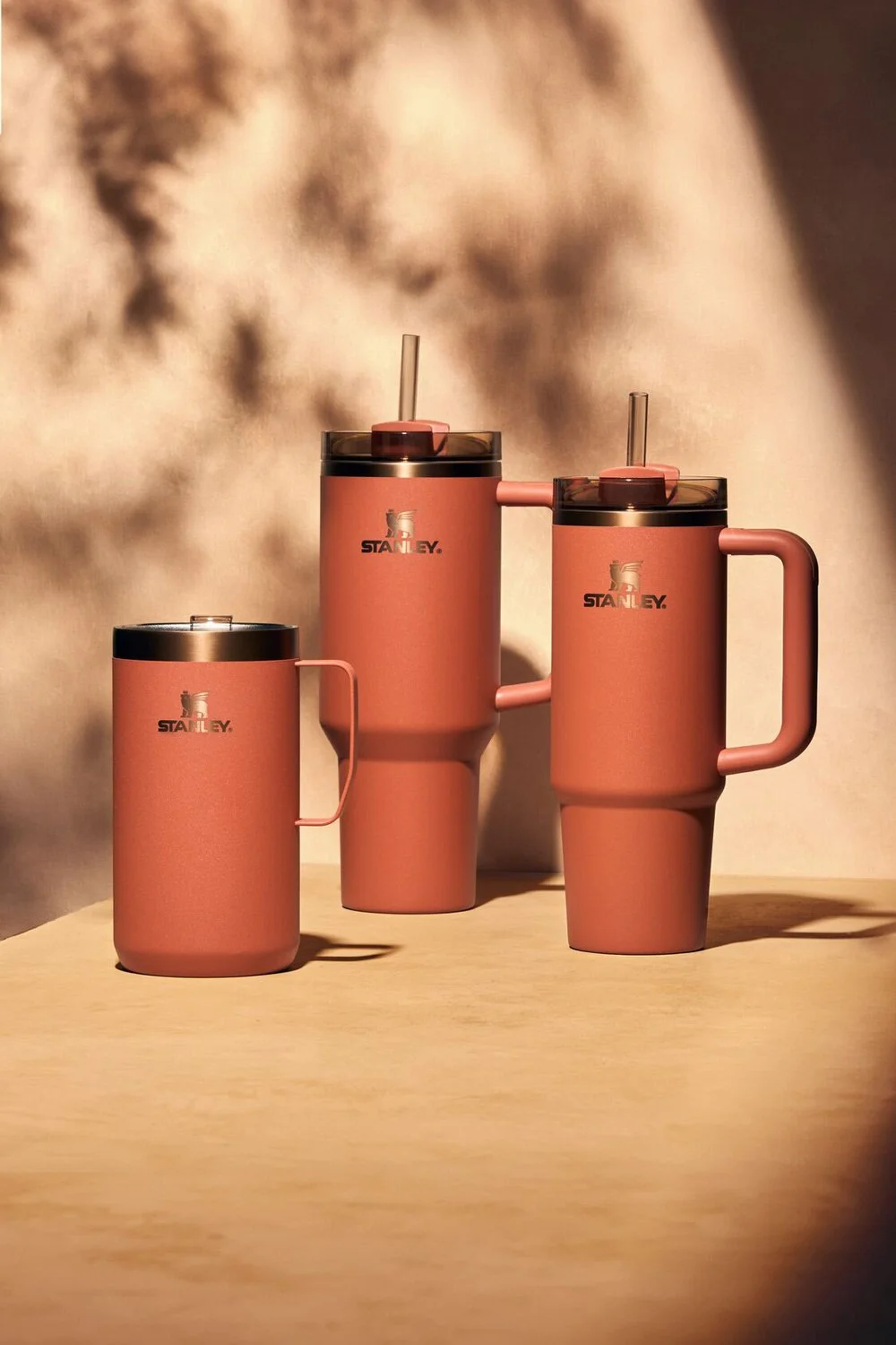 three pink Stanley insulated drink containers with black lids and straws, placed on a wooden surface, with a sandy background.