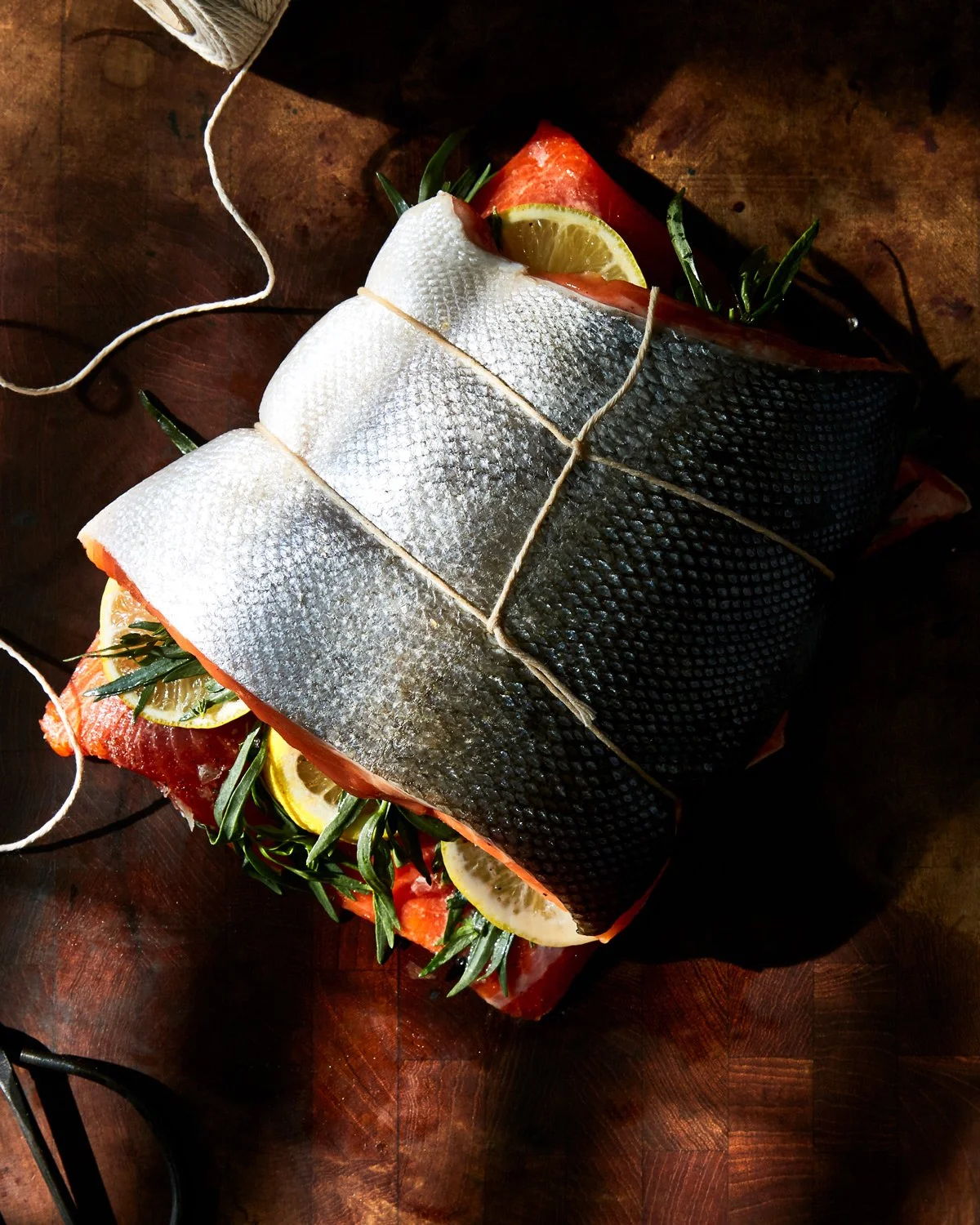 Raw fish fillet seasoned with lemon slices and herbs, secured with kitchen twine on a wooden surface.