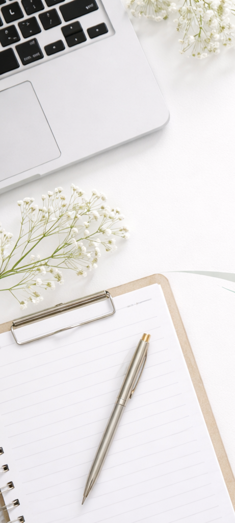 A silver pen resting on a spiral-bound clipboard with blank lined paper, white flowers, a silver laptop keyboard, and a white workspace surface.
