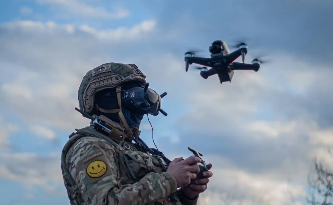 A soldier in camouflage uniform and tactical gear operates a drone with a remote control, against a backdrop of a cloudy sky.