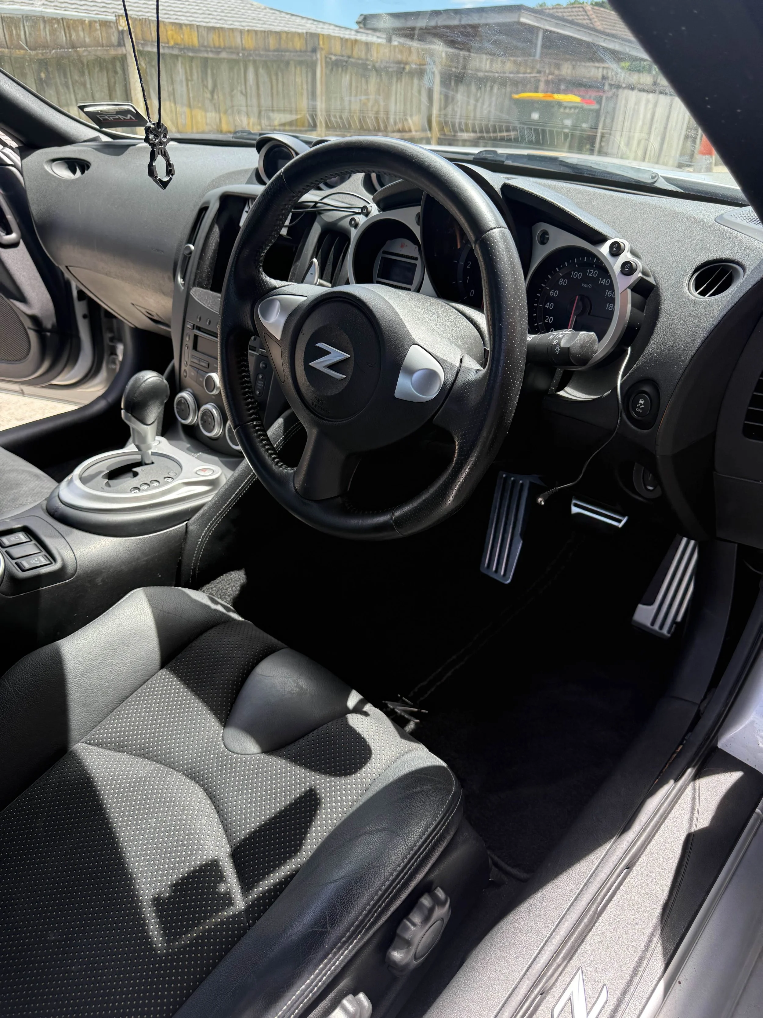 Inside the interior of a Nissan 370Z, showing the steering wheel, gear shift, dashboard, and driver's seat with shadow patterns, with an outside view through the windshield of a wooden fence and building.