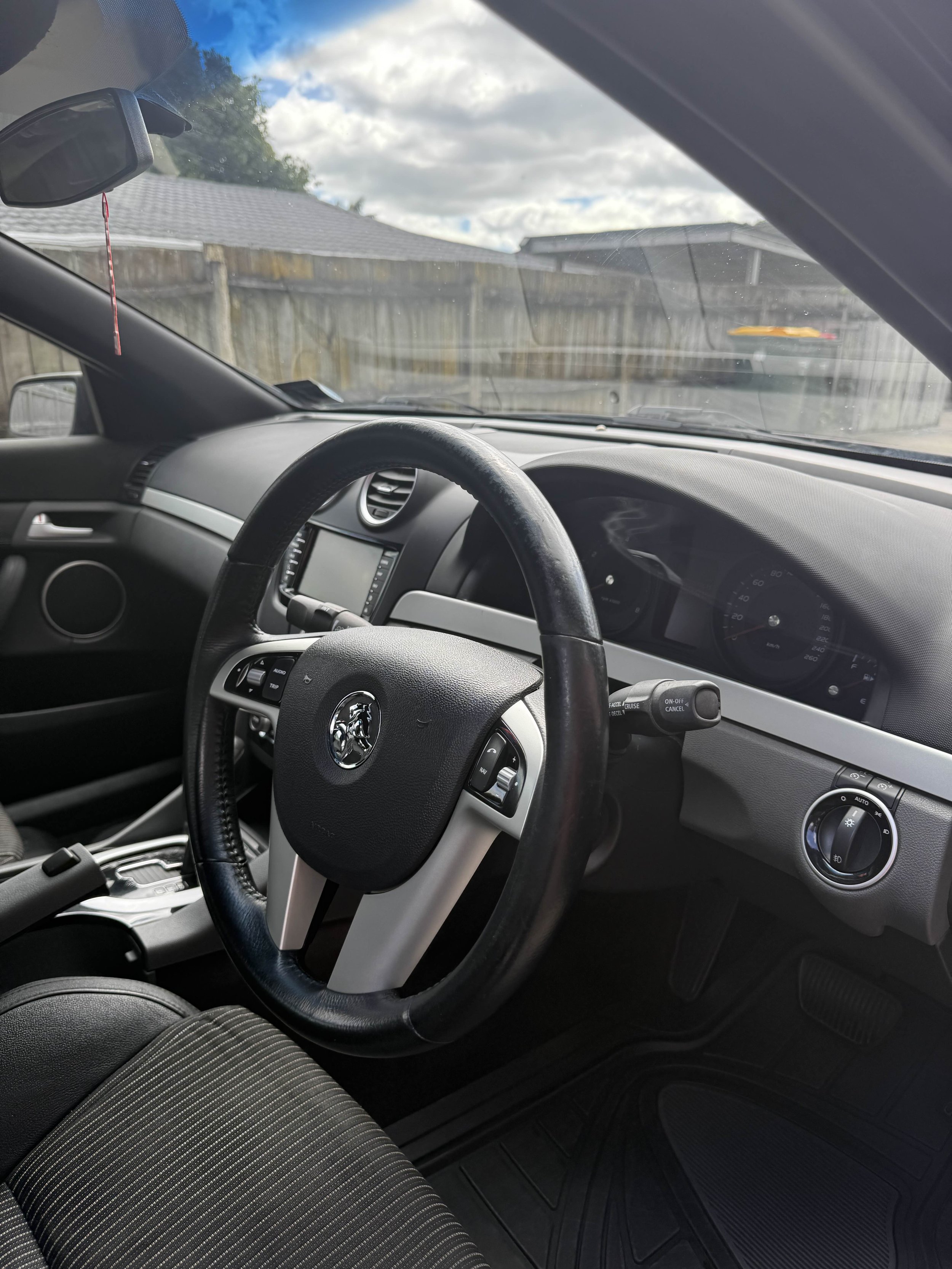 Interior view of a car showing the steering wheel, dashboard, and driver's seat, with an outside view of a backyard with a wooden fence and cloudy sky.
