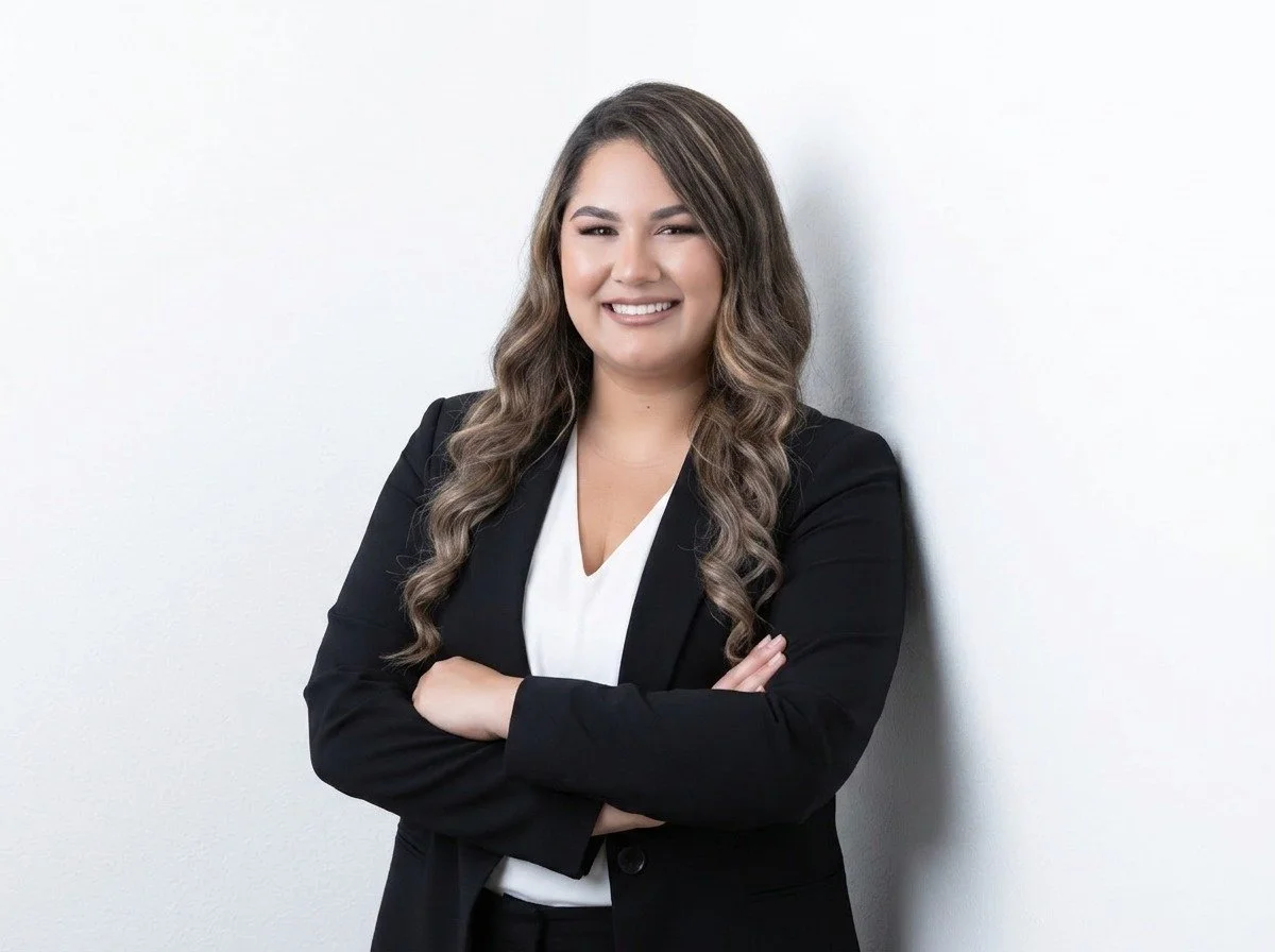 A woman smiling and winking while standing with arms crossed against a white wall, dressed in a black blazer and white blouse.