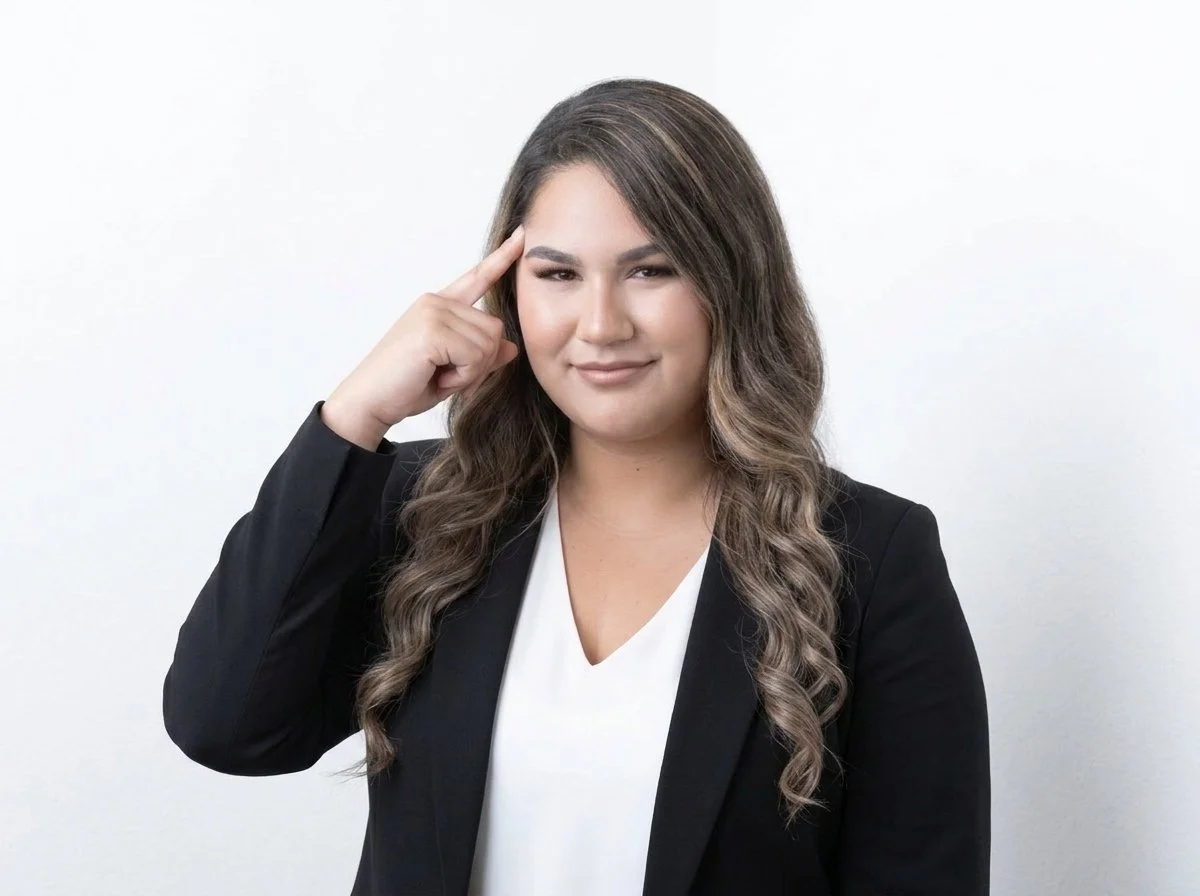 A woman with wavy brown hair pointing to her temple, wearing a black blazer over a white top, against a plain white background.