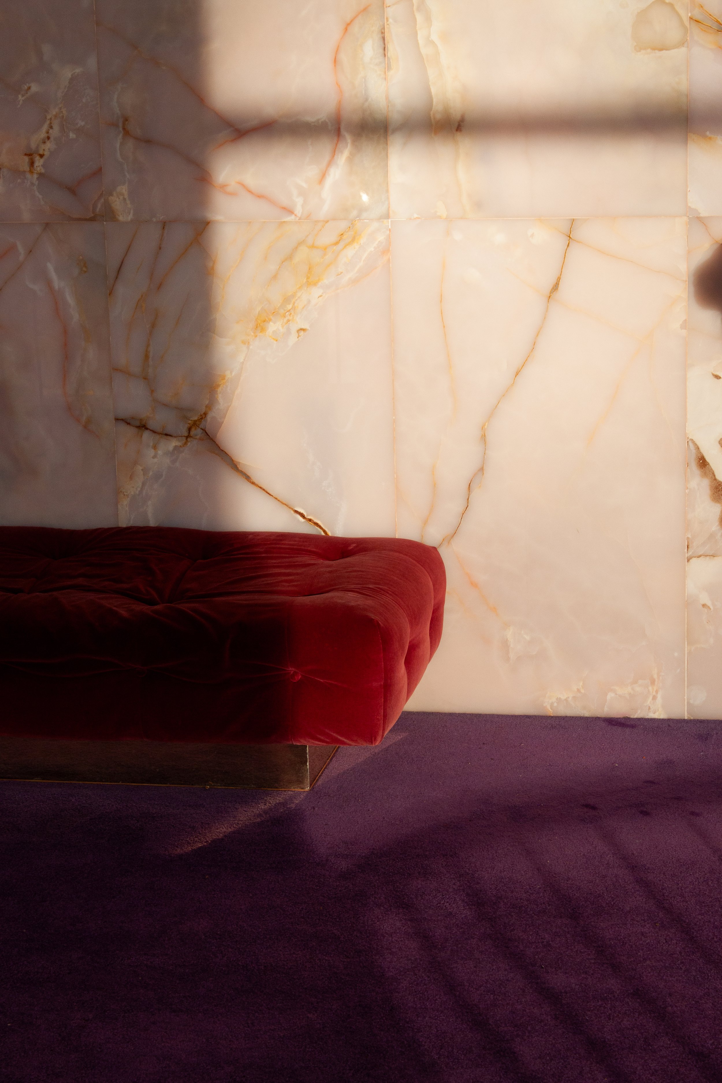 A plush red velvet sofa with deep button tufting, positioned on a purple carpet in front of a large, illuminated onyx wall with orange and beige veining.