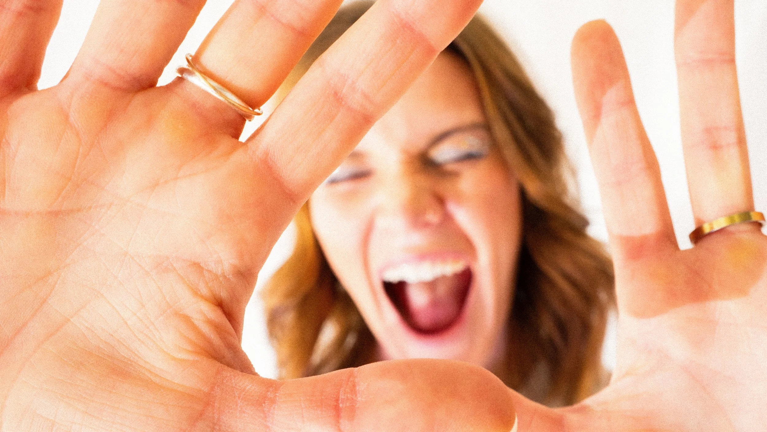 Close-up of a woman's face with her mouth open, hands in front making a frame, with rings on her fingers, and a blurred background.