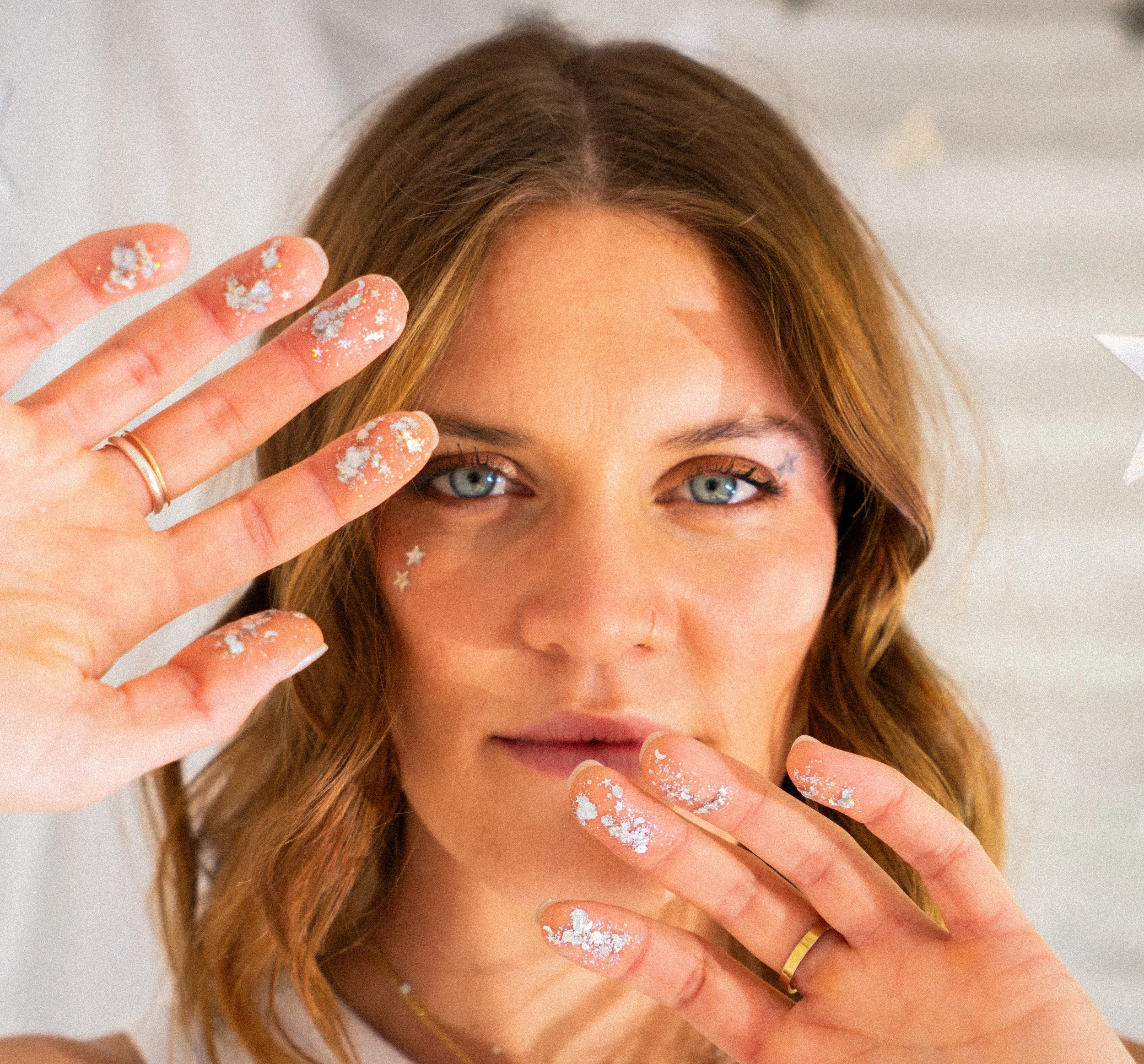 A woman with light brown hair and blue eyes, holding her hands near her face, which are covered in glitter and star-shaped confetti.