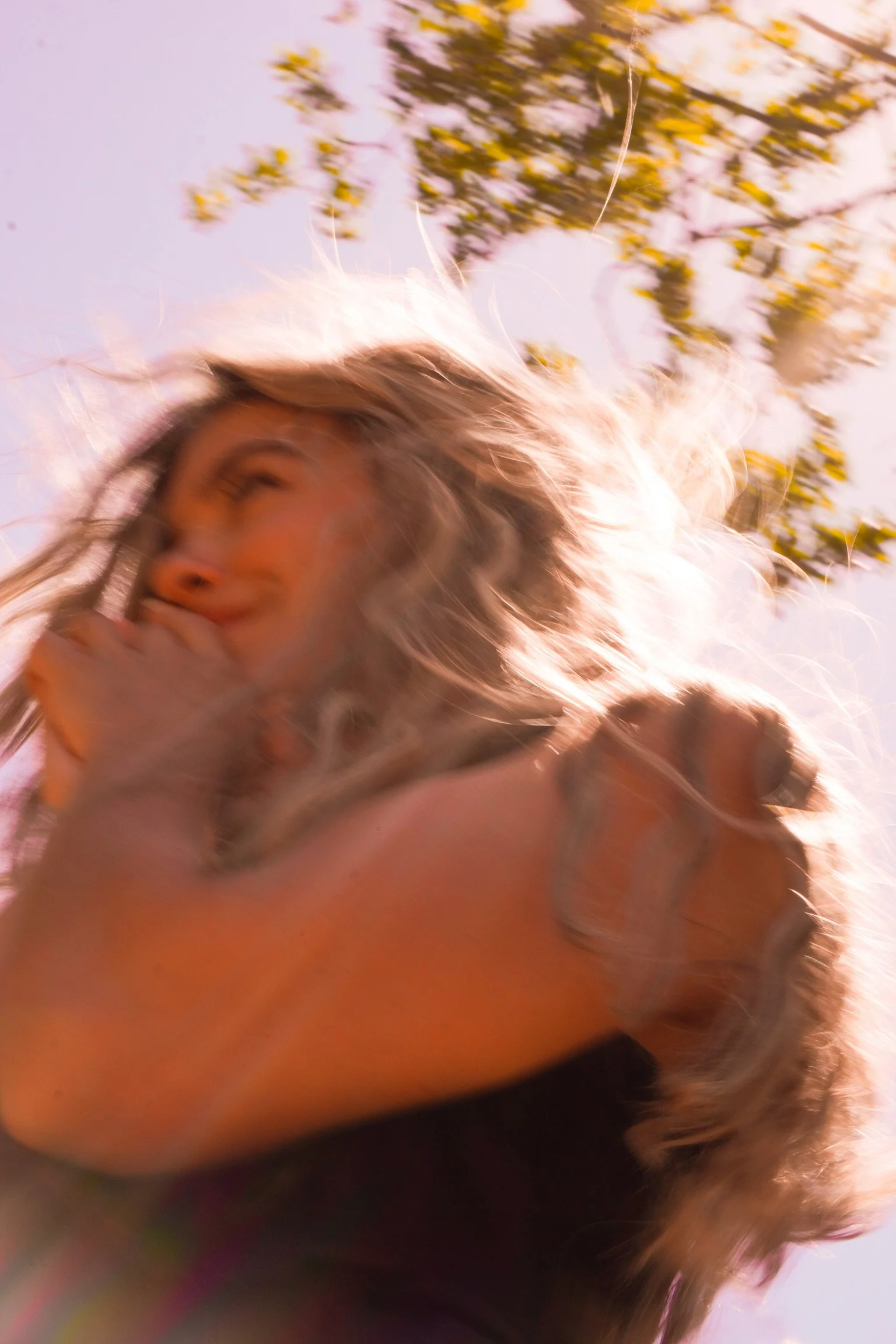 A person with long, wavy hair outdoors, smiling with eyes closed, sunlight illuminating their face and hair, against a background of tree branches and sky.