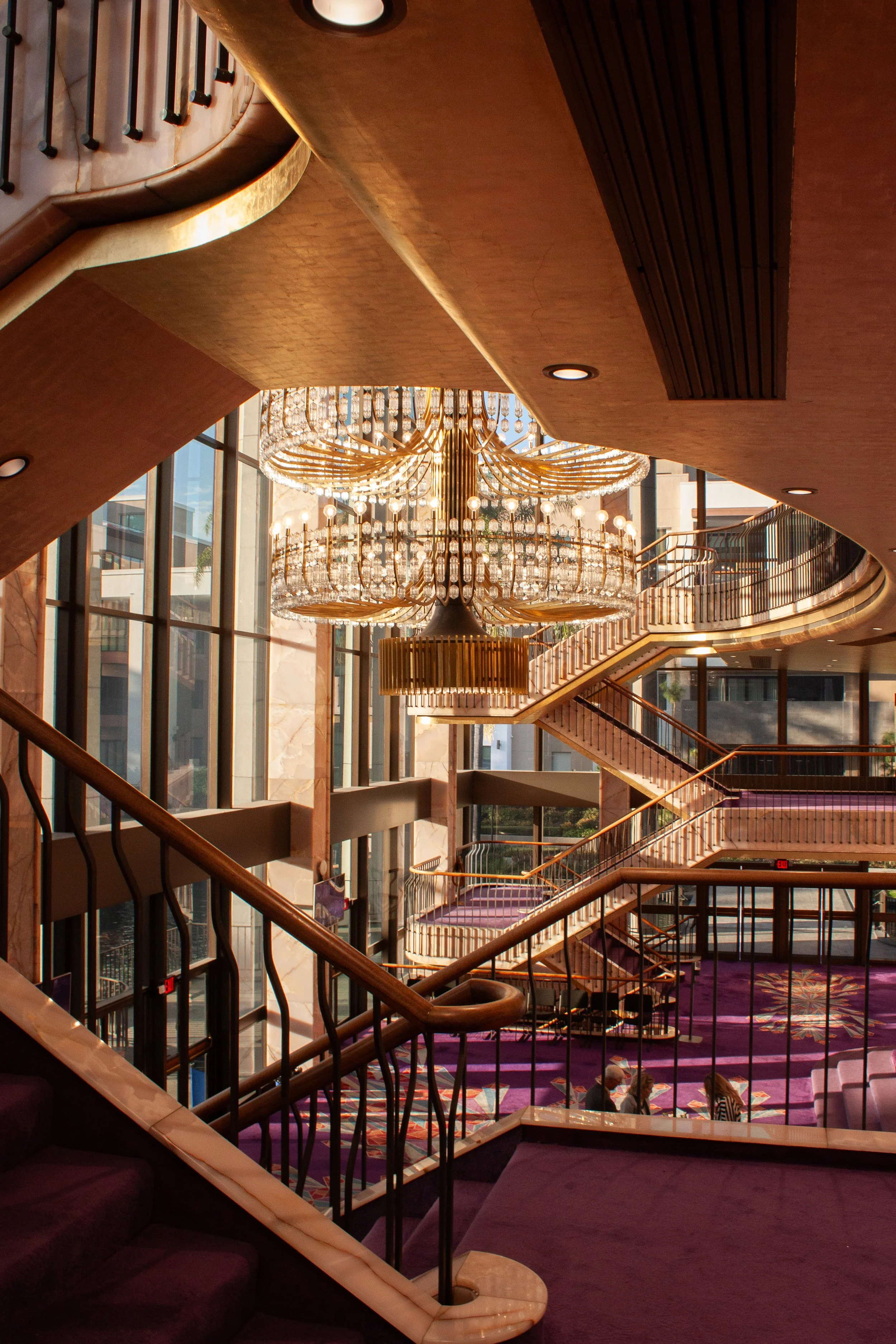 Interior of a hotel lobby with a large crystal chandelier hanging from the ceiling, multiple staircases with wooden handrails, and purple carpeted floors.