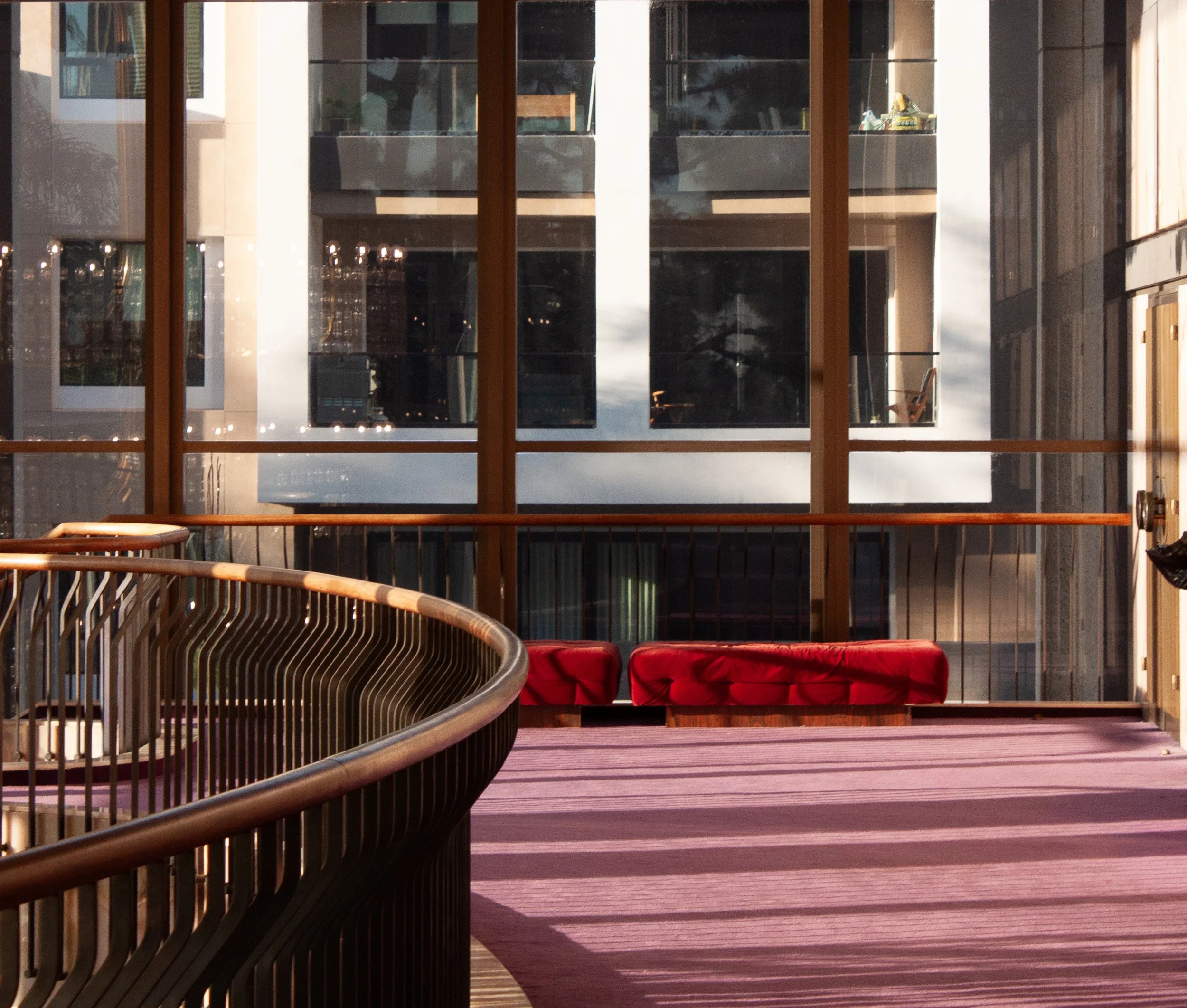 Interior view of a modern building with large glass windows, a curved wooden railing, a purple carpet, and a red bench.