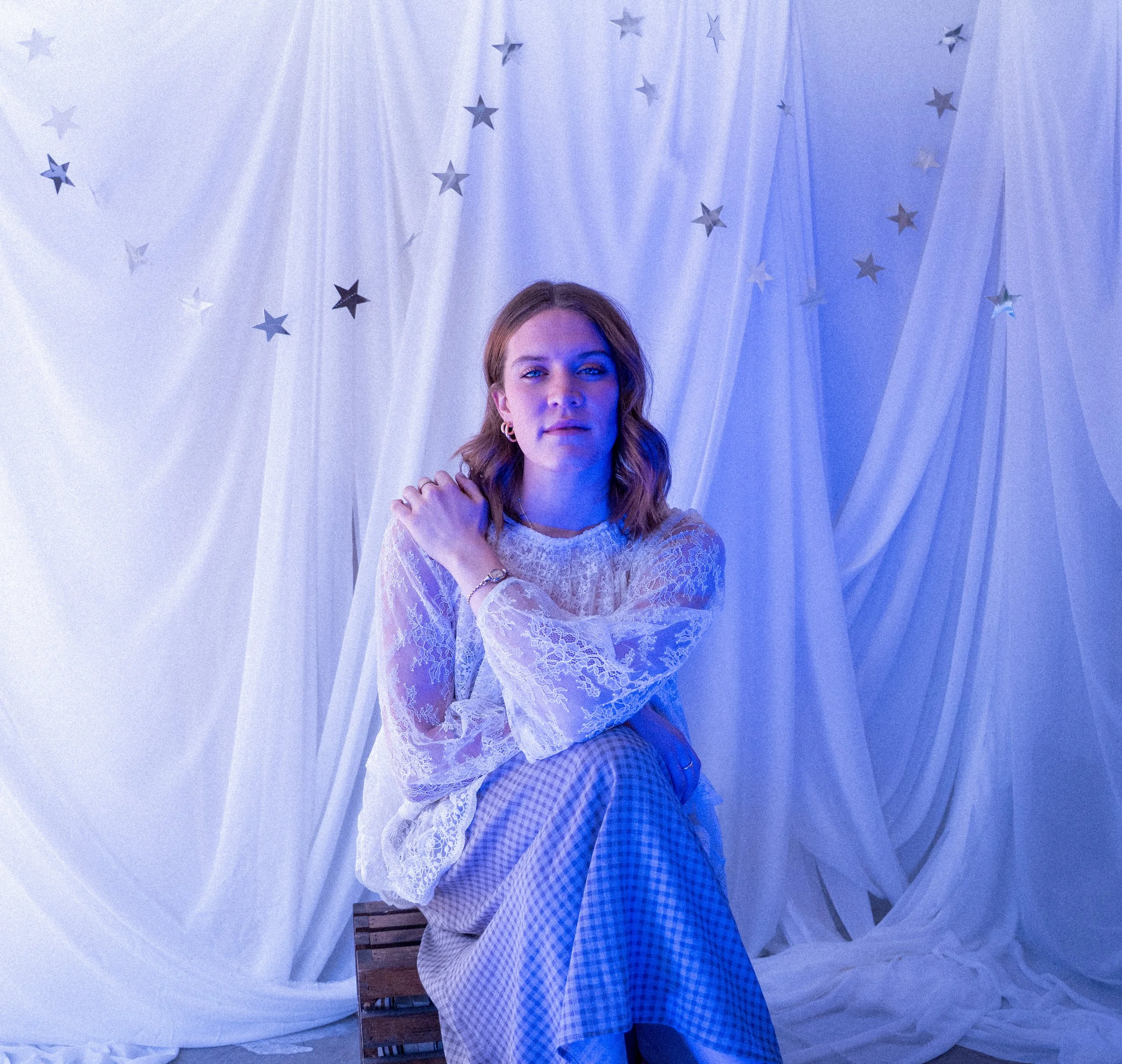 A woman sits on a wooden crate in front of a backdrop of white drapes decorated with silver stars, illuminated with blue lighting.