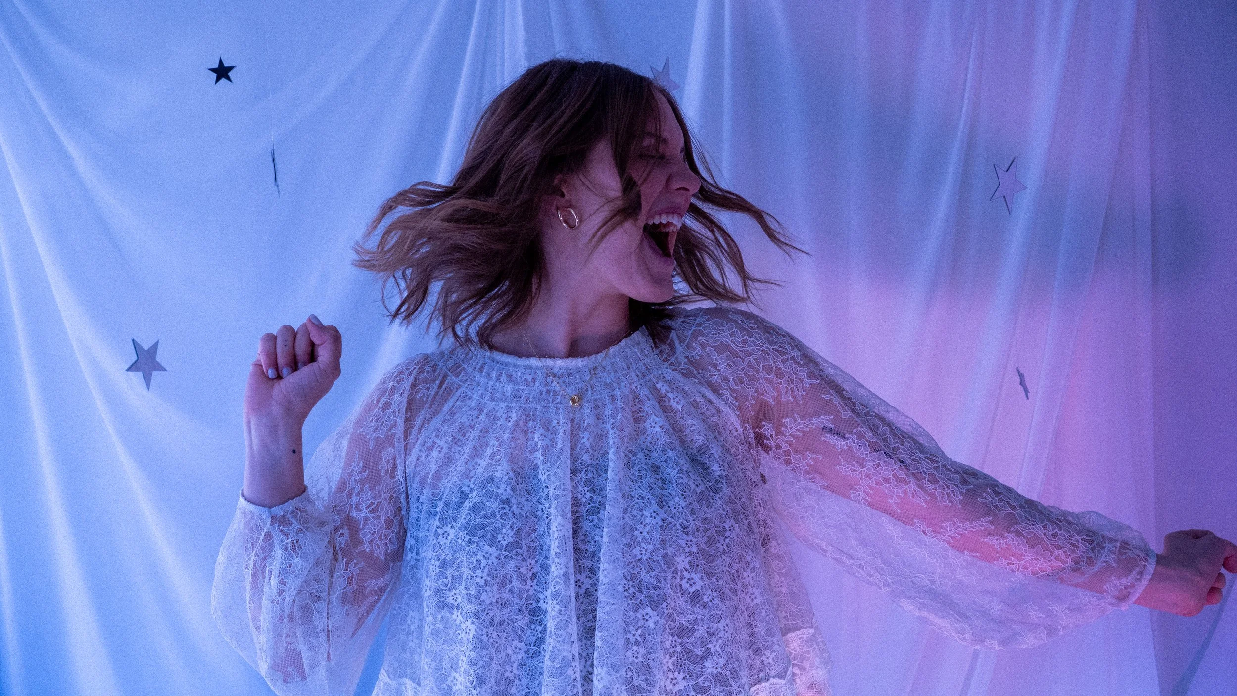 A woman with short brown hair, wearing a lacy white dress, is dancing and singing with her mouth open and her eyes closed. The background features a white curtain with star-shaped decorations and colorful lighting.