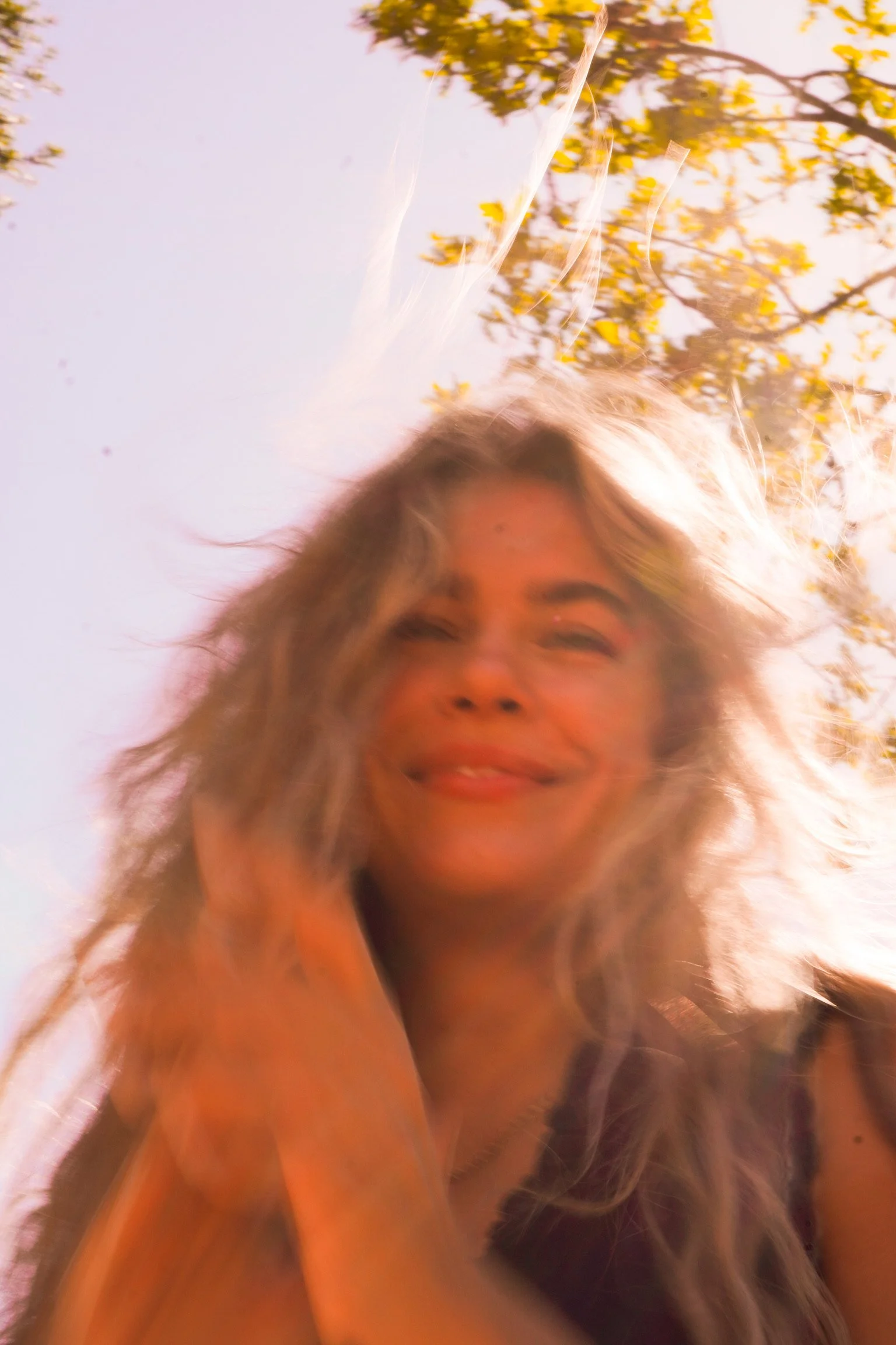 A woman outdoors with sunlight and trees in the background, her hair blown by the wind, smiling.