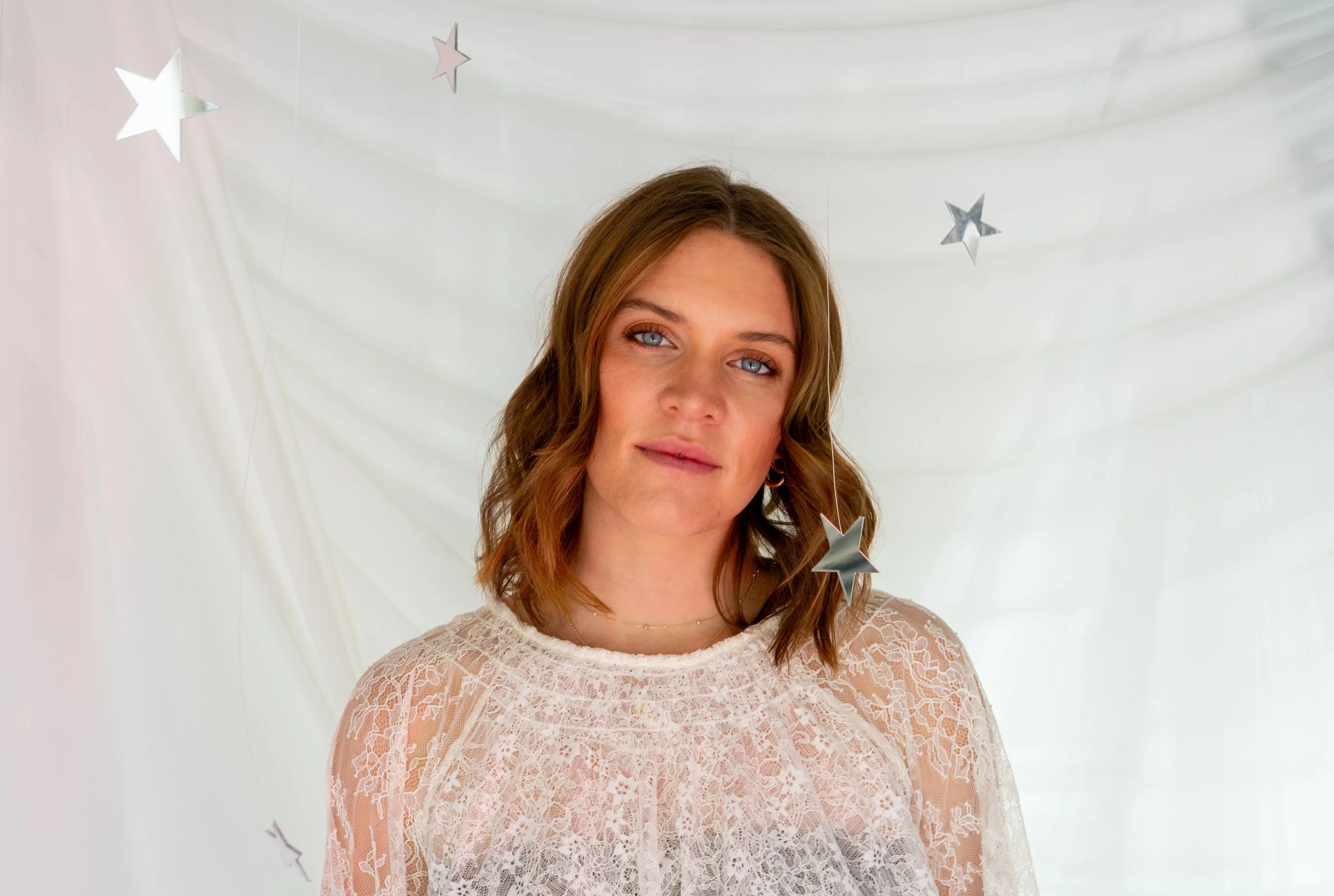 A woman with shoulder-length brown hair, blue eyes, and light skin, wearing a white lace top, standing in front of a white backdrop decorated with hanging silver and pink stars.