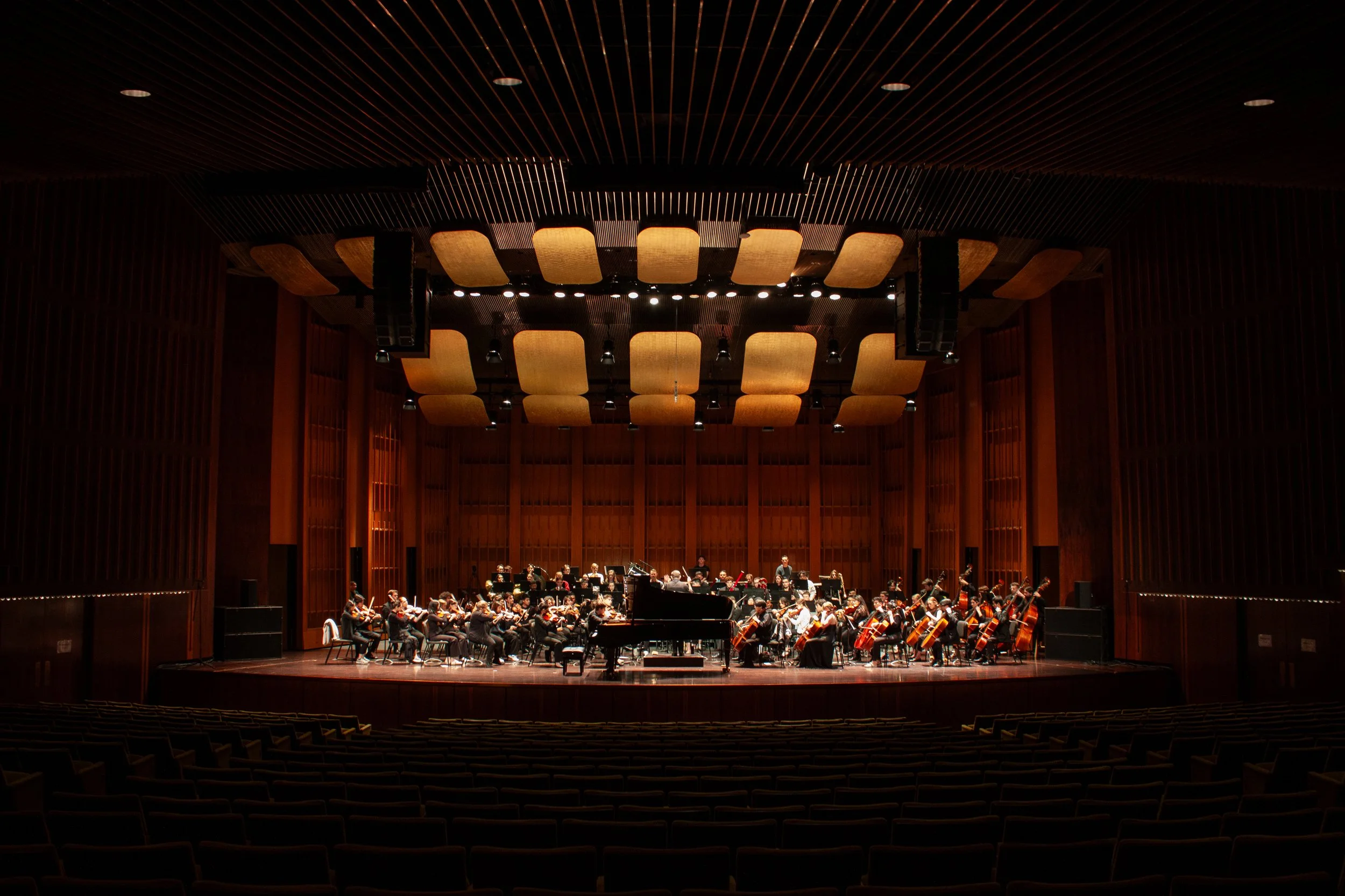 An orchestra performing on a large wooden stage inside a concert hall with warm lighting, surrounded by empty seats.
