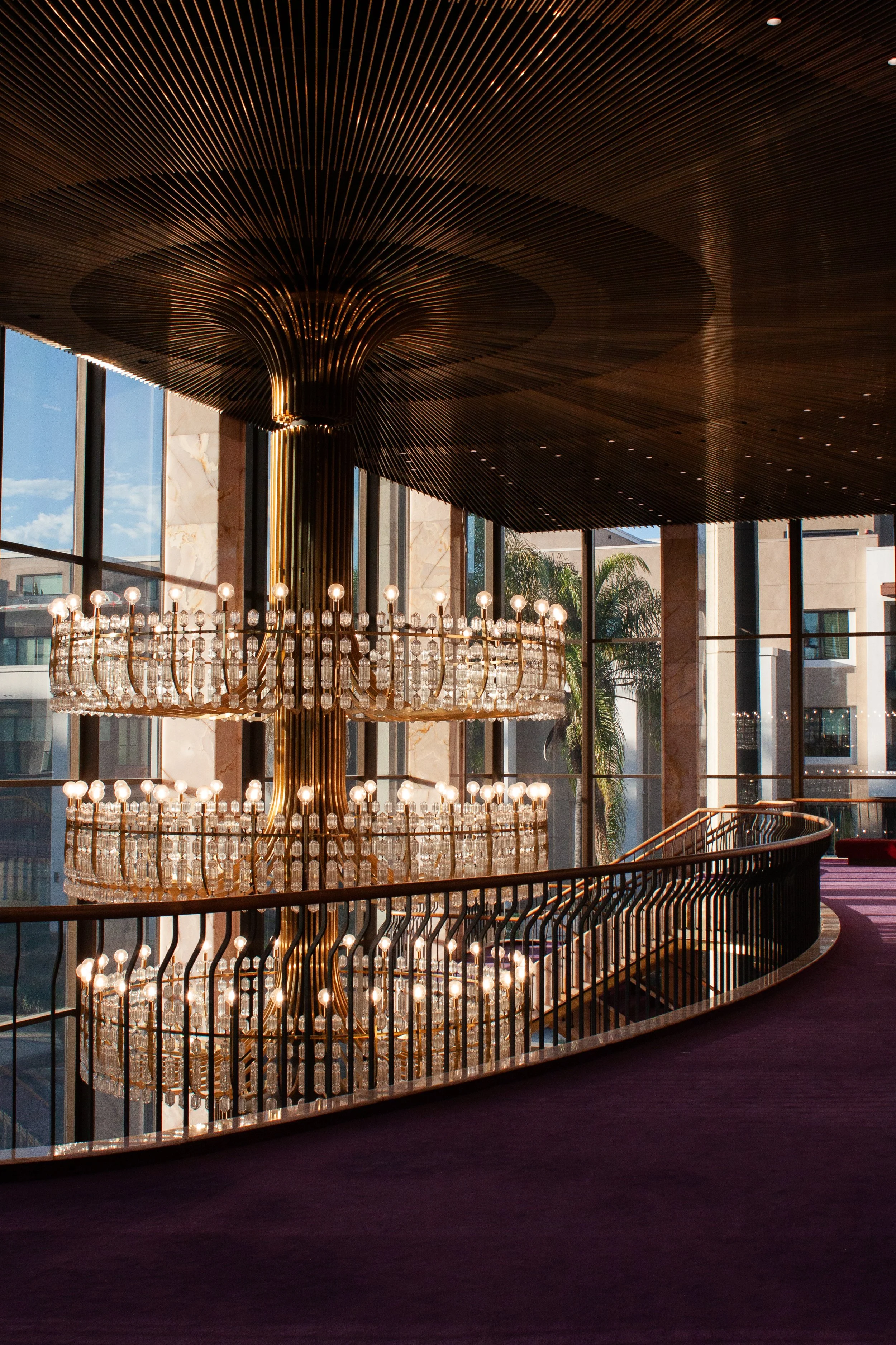 Elegant interior with a large, multi-tiered crystal chandelier, curved balcony railing, tall windows with a cityscape background, and a wooden ceiling with radiating slats.